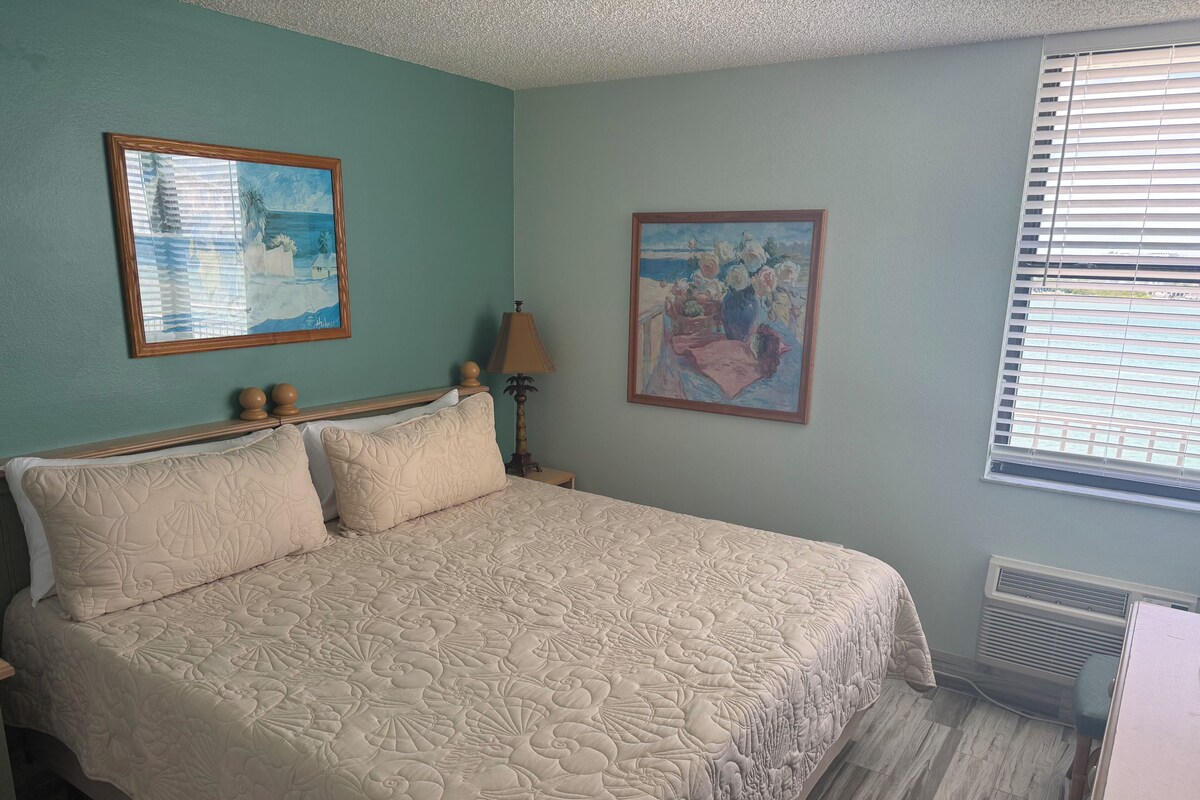 A bed with a floral-patterned bedspread, a nightstand with a lamp, and two framed artworks on the walls.