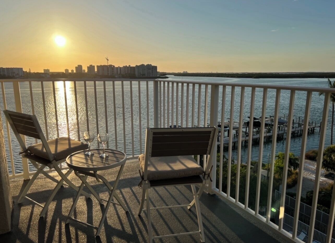 A balcony with two chairs and a table, overlooking a waterfront with buildings and a boat.