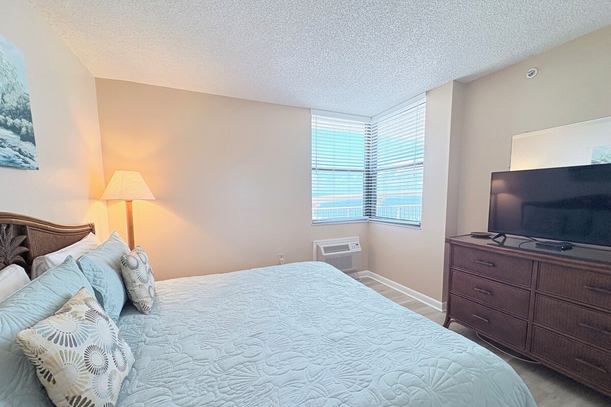 A bedroom with a bed, a TV, a dresser, and a window with blinds.