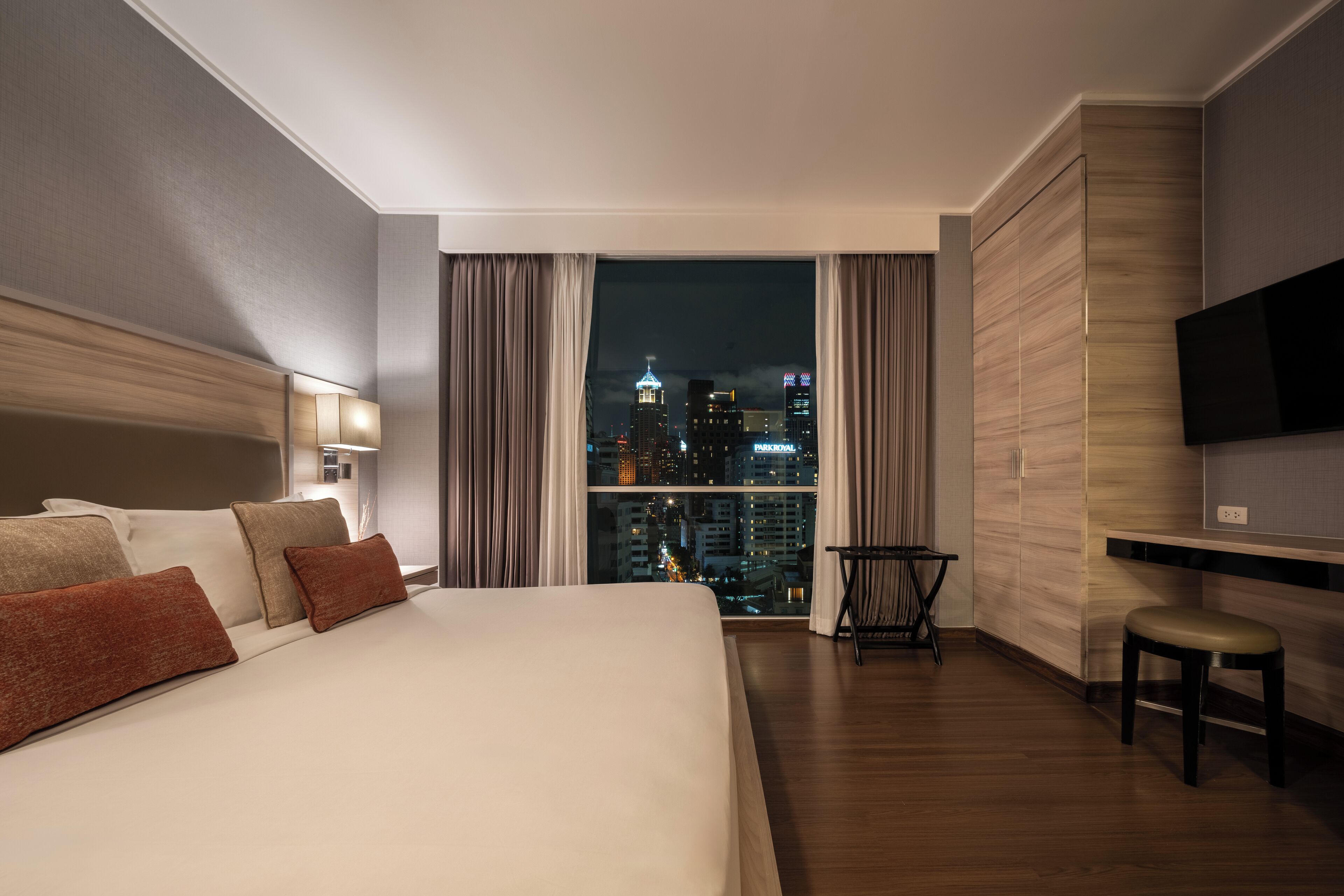 A hotel room with a large bed, a TV, a desk, and a view of a city skyline at night.