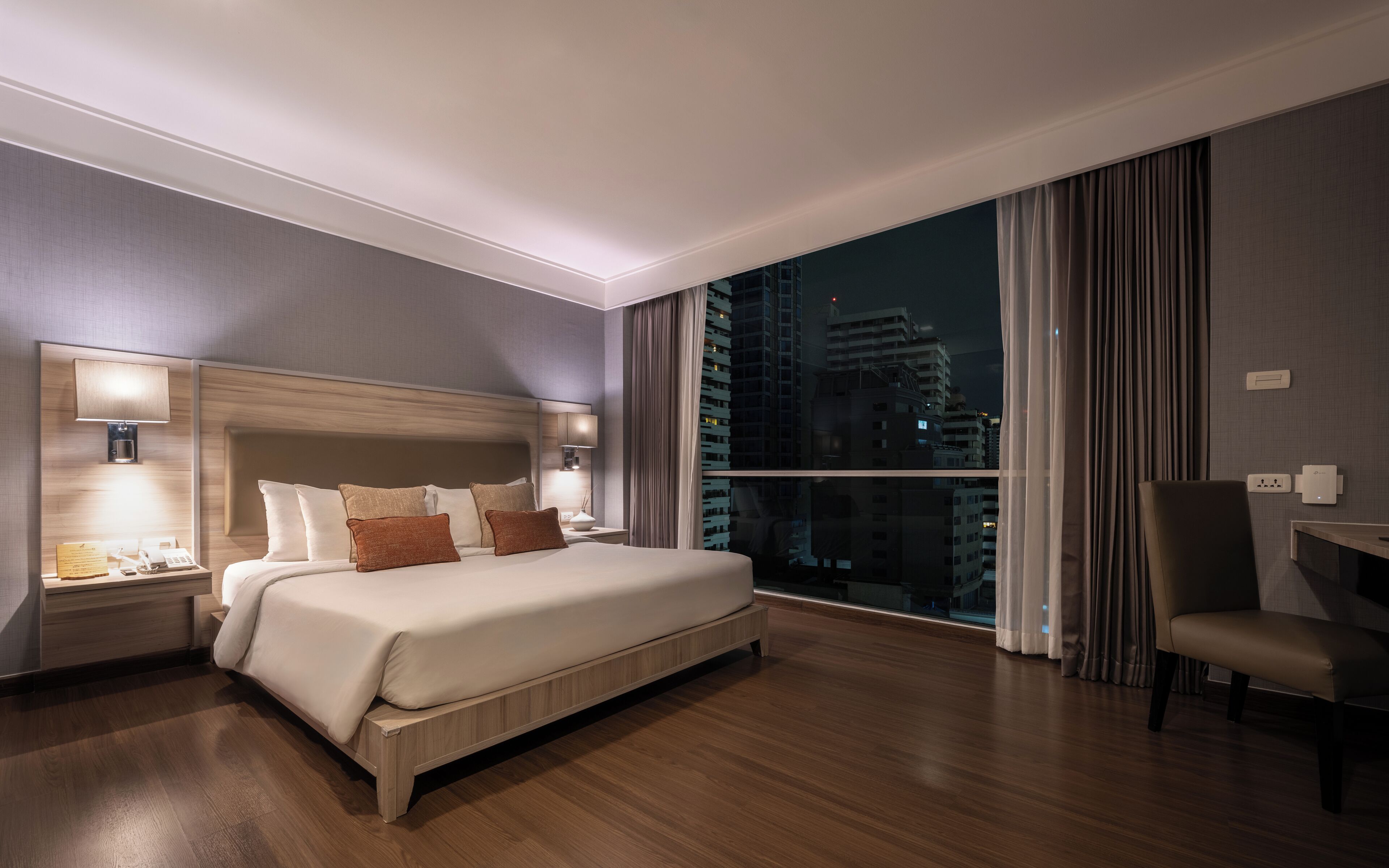 A modern hotel room with a large bed, bedside lamps, a desk, and a view of the city at night.