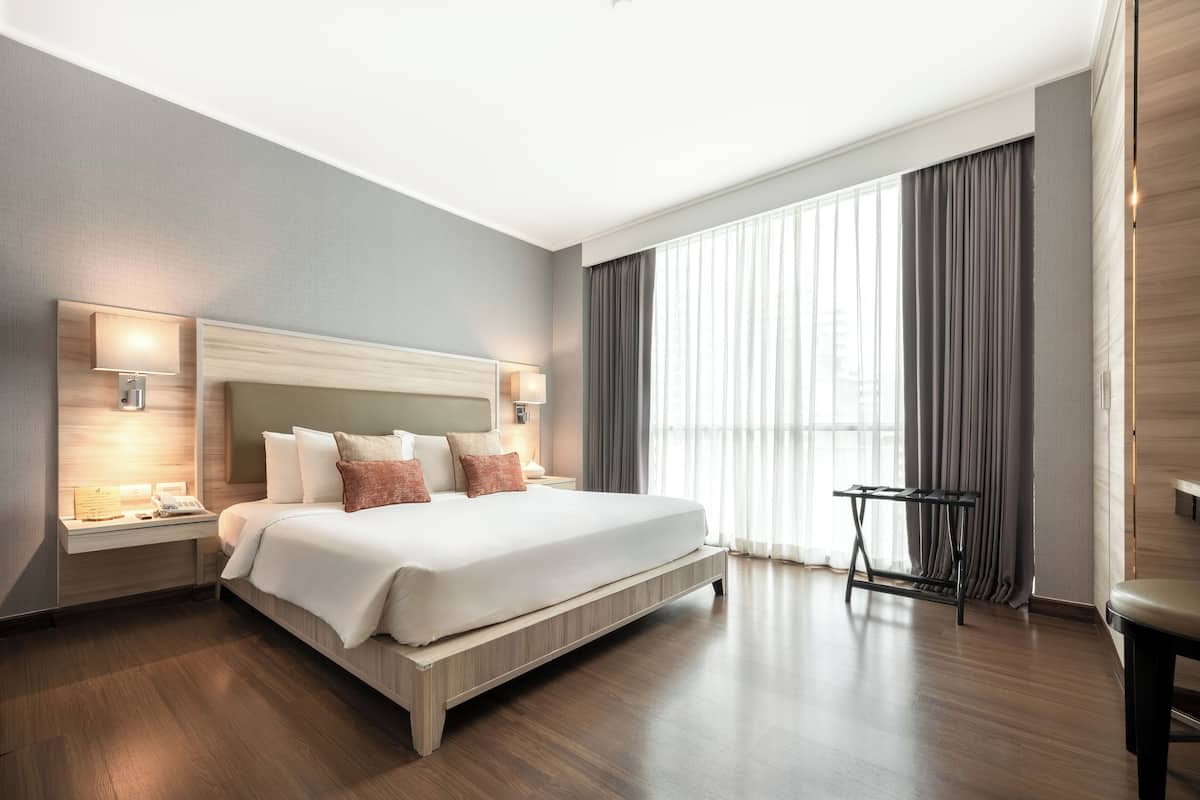 A modern hotel room with a large bed, bedside lamps, a small table, and a view of the outdoors through large windows.