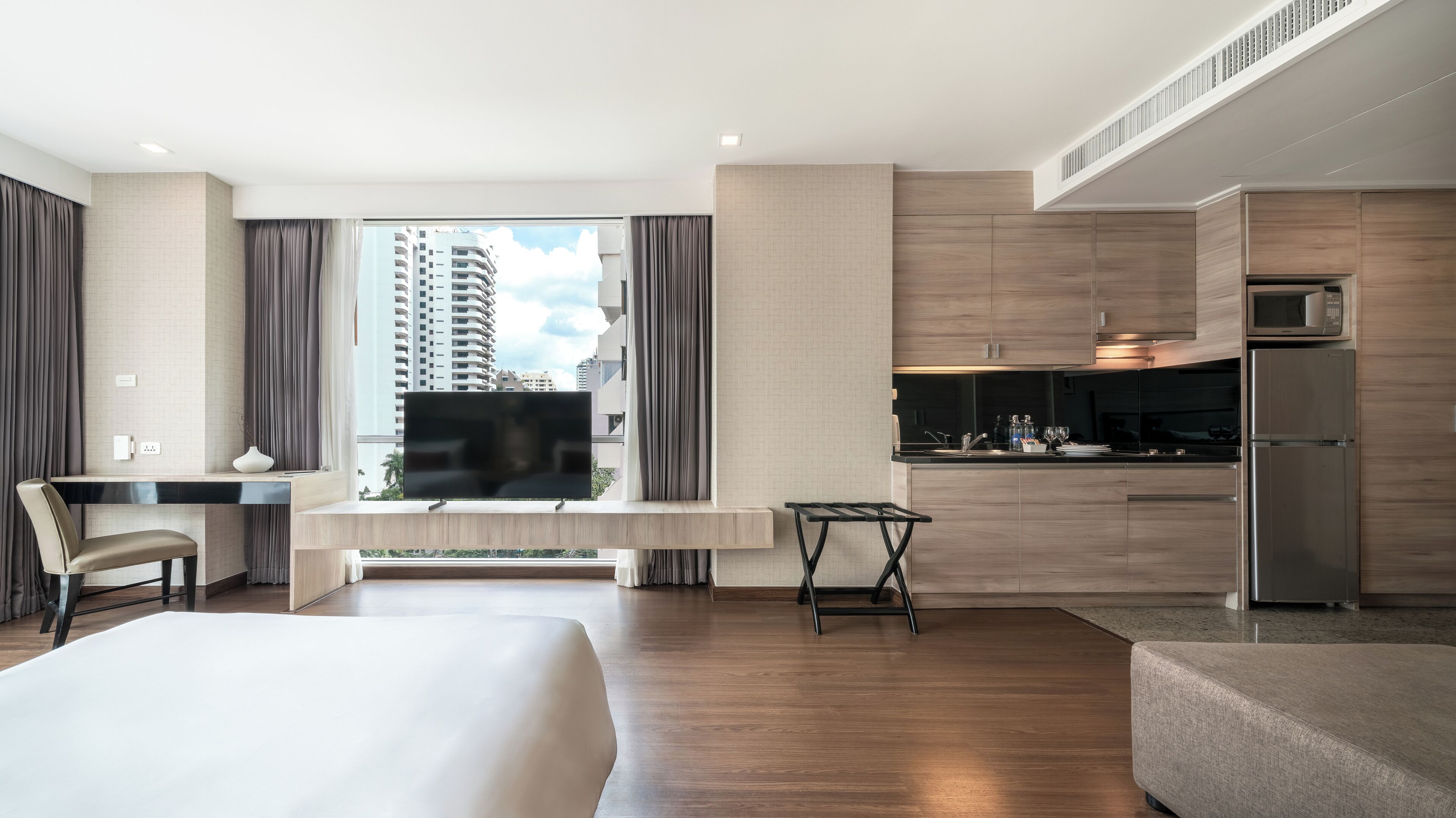 A modern hotel room with a large bed, a desk, a chair, a small table, a sofa, a kitchenette with a microwave and refrigerator, and a view of tall buildings.