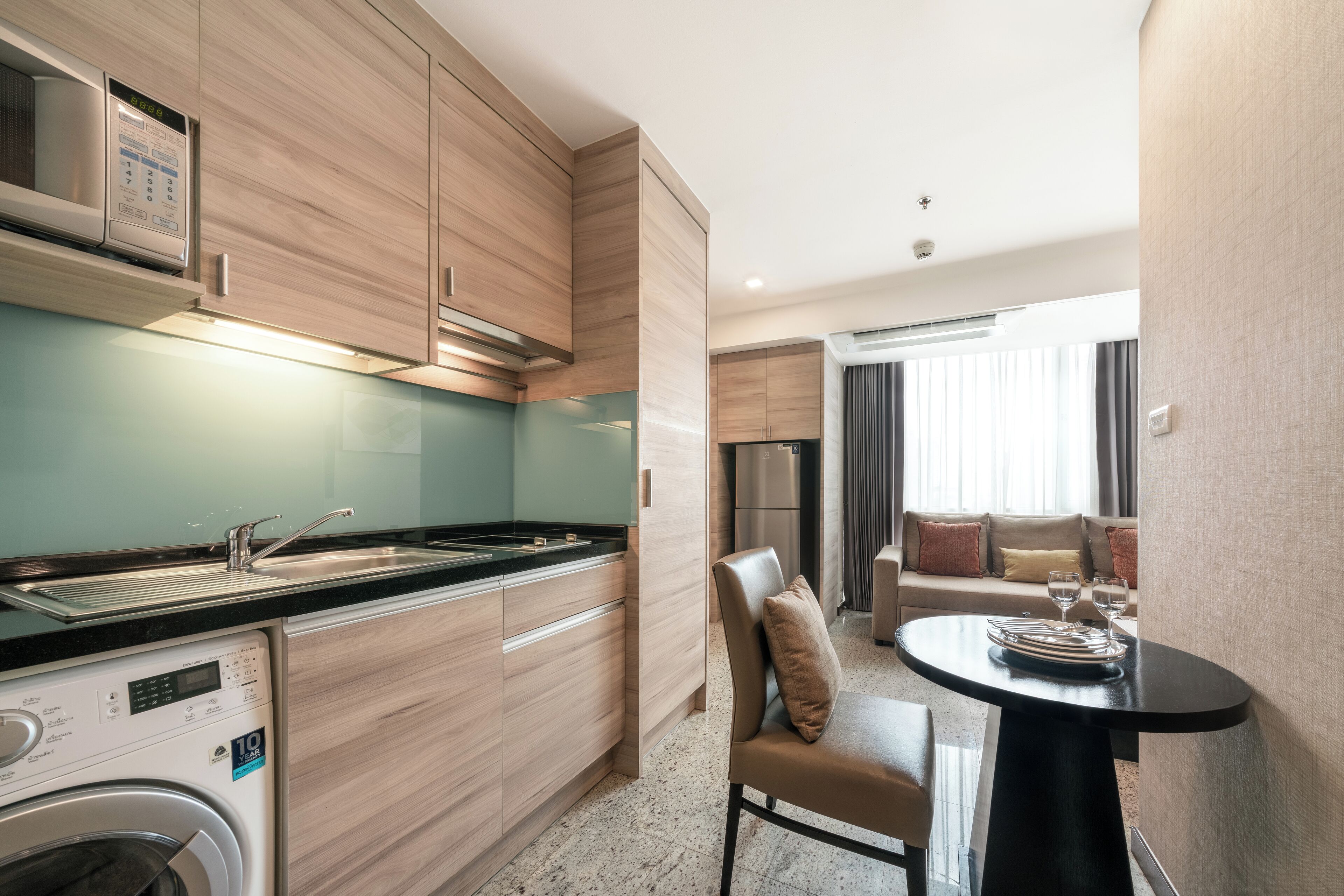 A modern kitchen with wooden cabinets, a built-in microwave, and a washing machine. There is a dining area with a round table and a sofa.