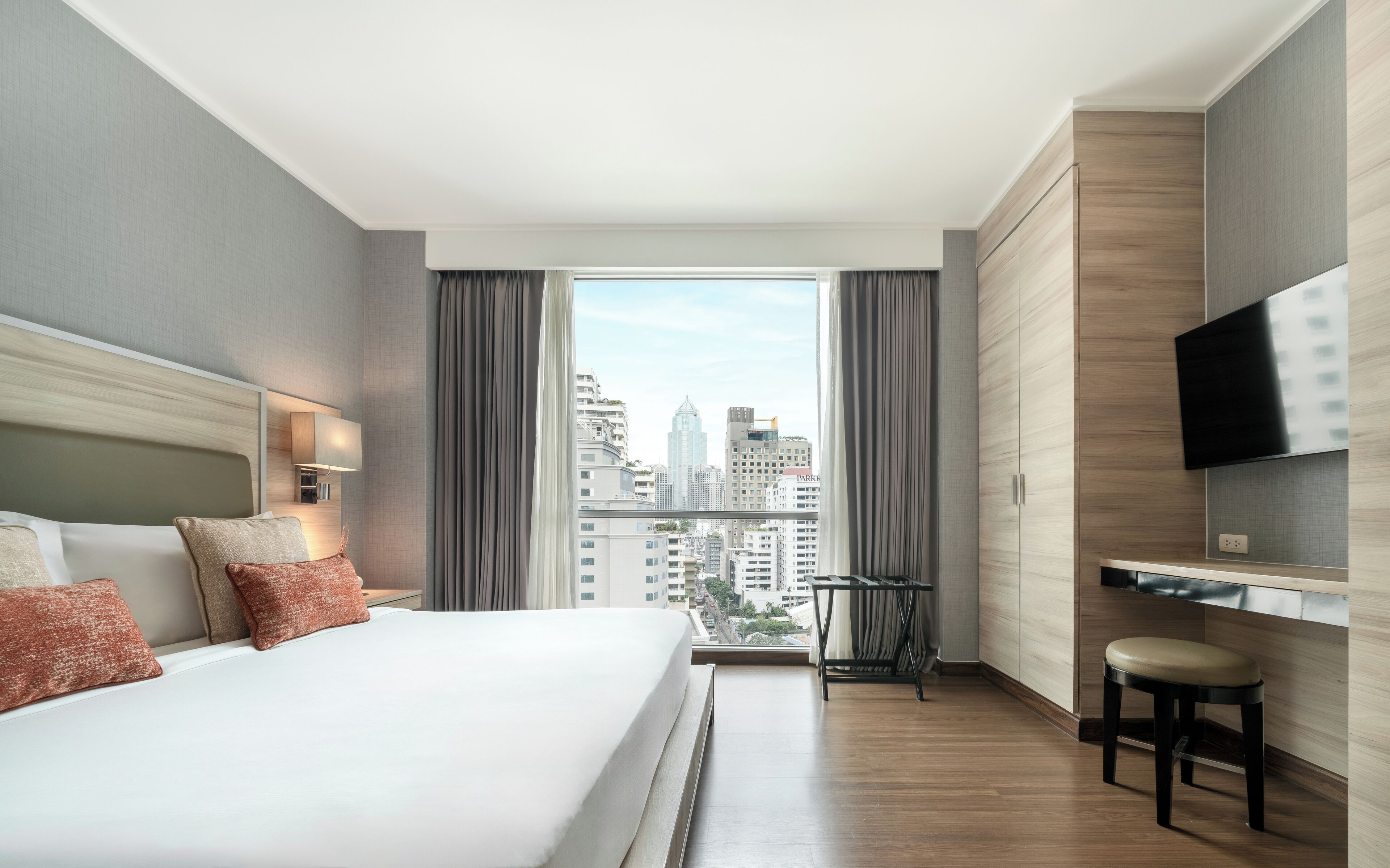 A modern hotel room with a large bed, a TV mounted on the wall, and a city view through the window.