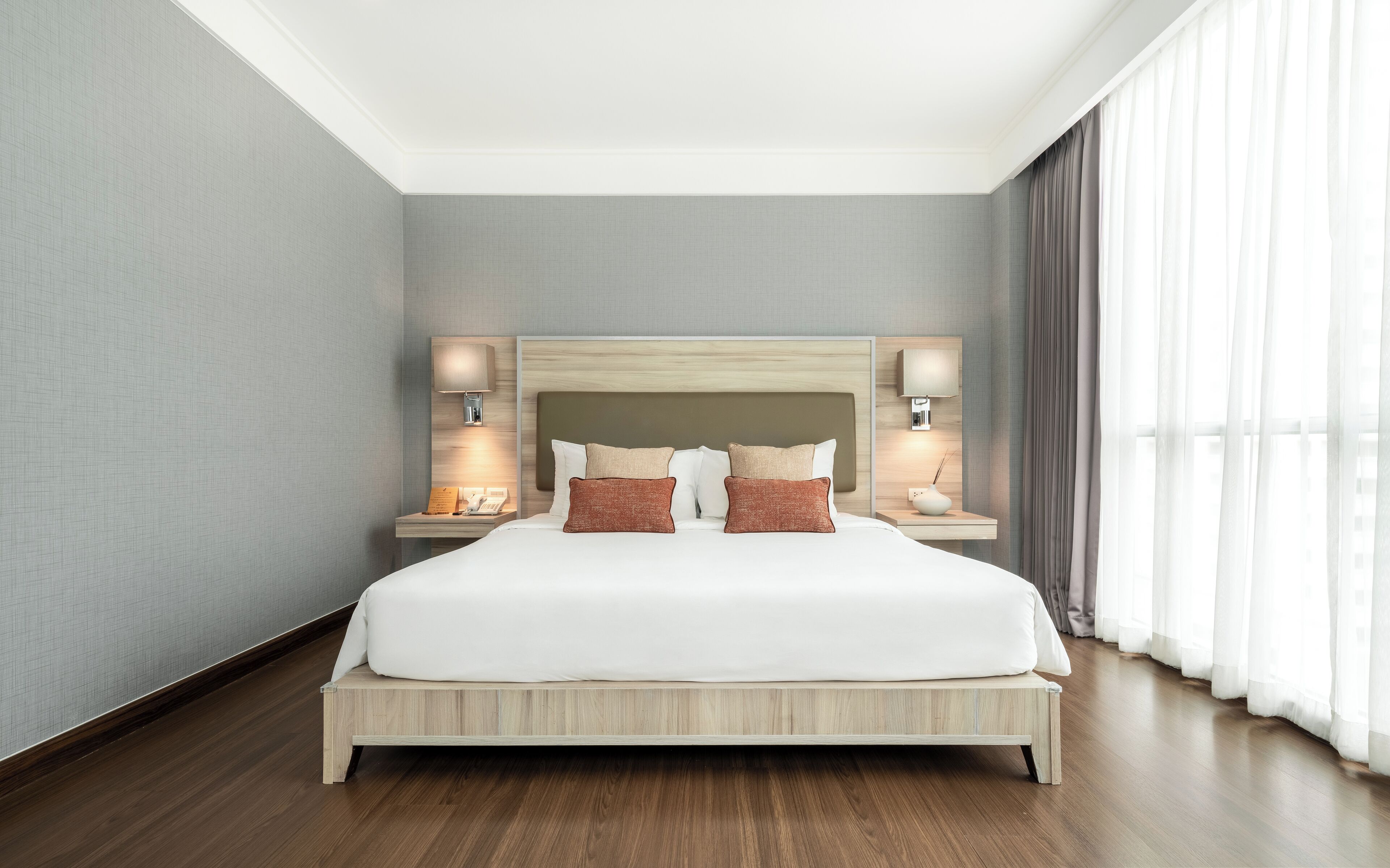 A modern hotel room with a large bed, wooden headboard, bedside lamps, and a view of the city through sheer curtains.