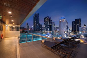 A rooftop pool area with lounge chairs and a city skyline view at dusk.