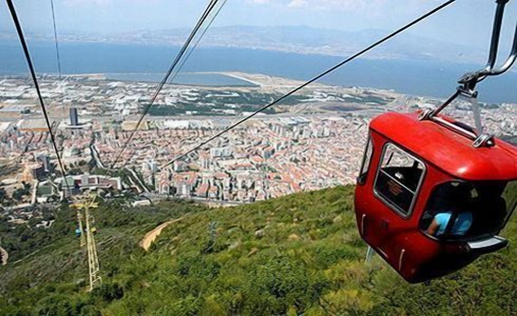 Una funivia con esterno rosso, sospesa da cavi, che offre vista su una città costiera con edifici e una torre imponente.