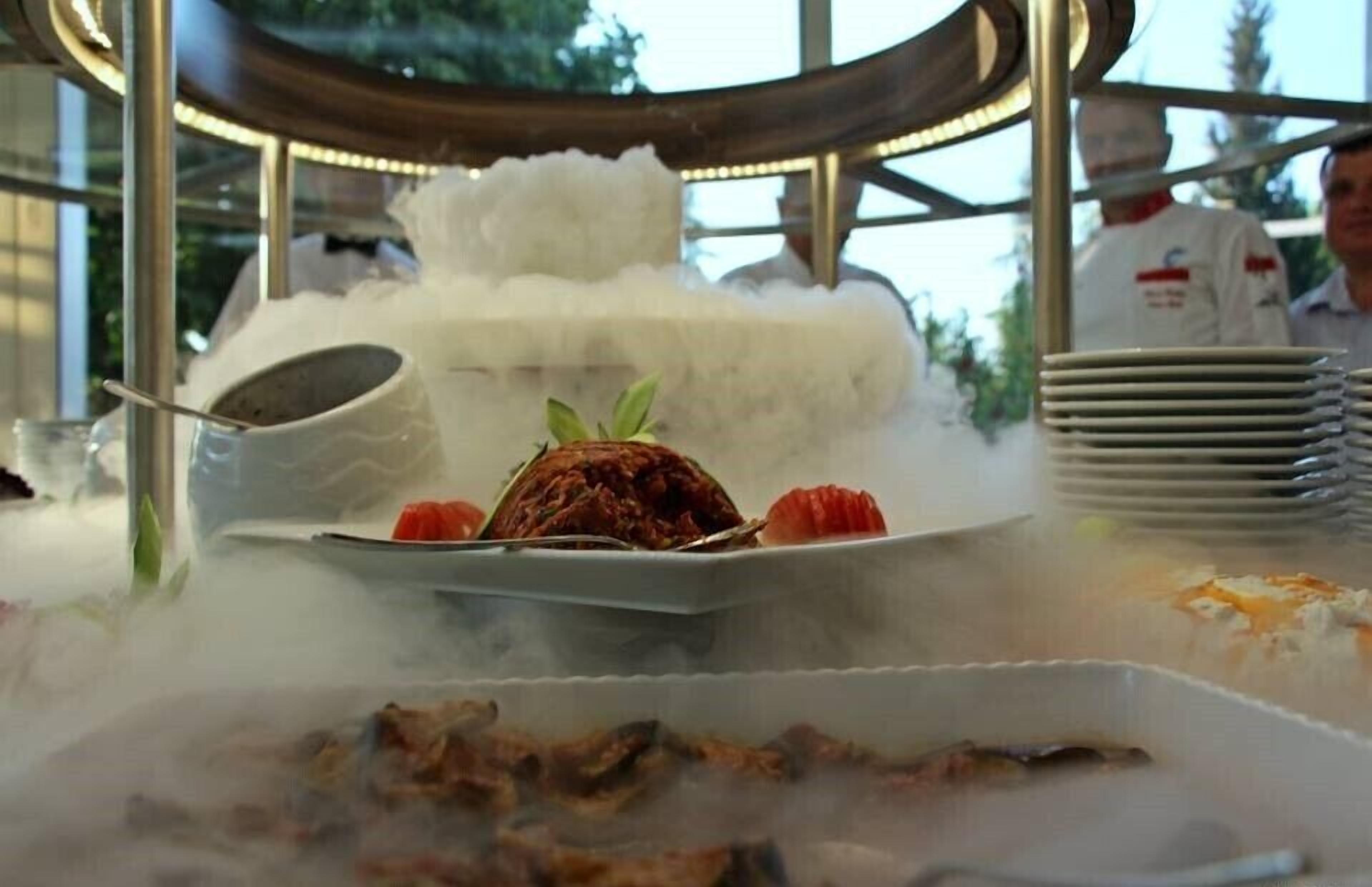 A dessert display with a cake and fruits, surrounded by steam, on a table with plates and a metal stand.