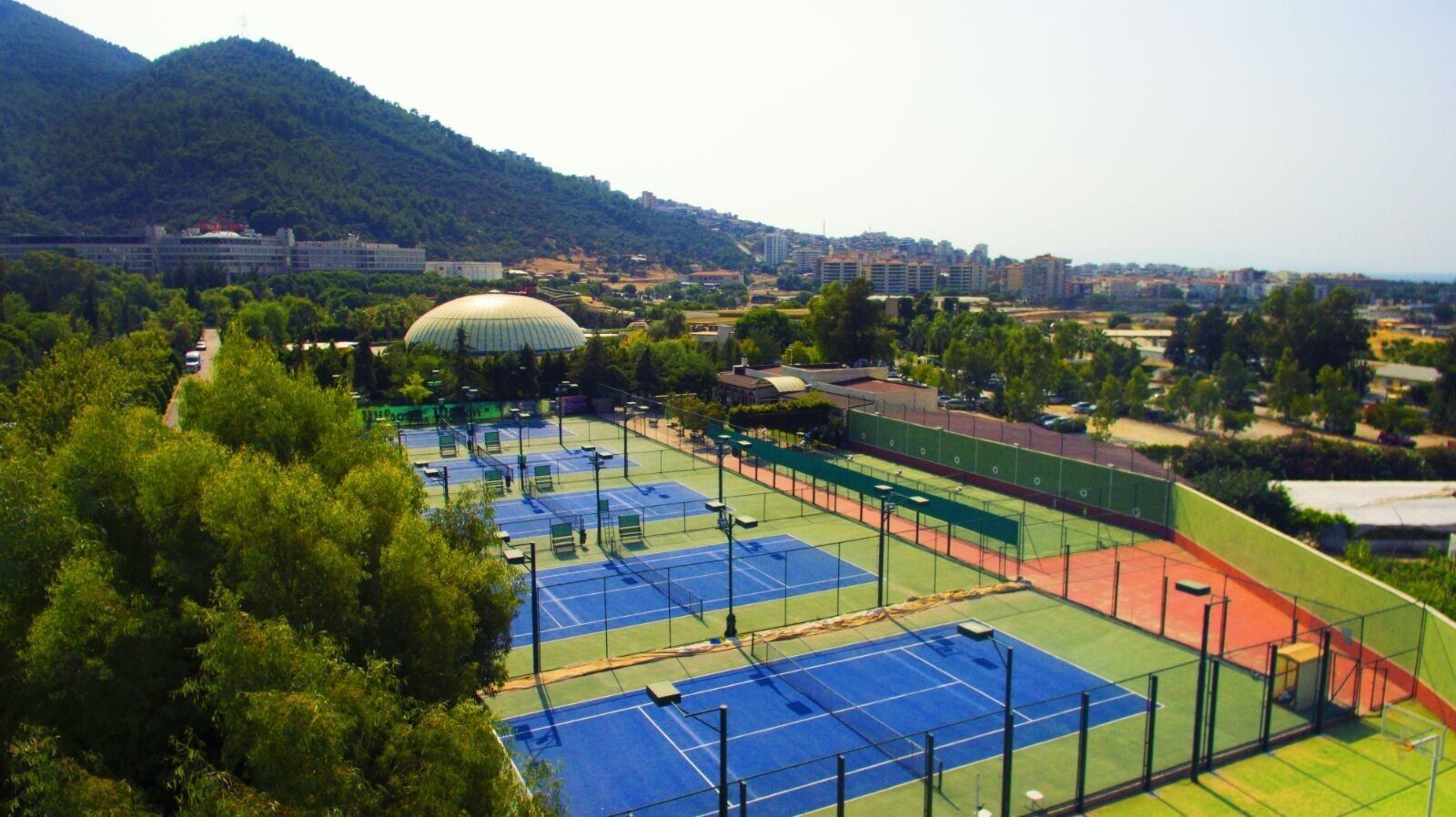 A tennis court complex with multiple courts, surrounded by greenery and a mountainous backdrop.