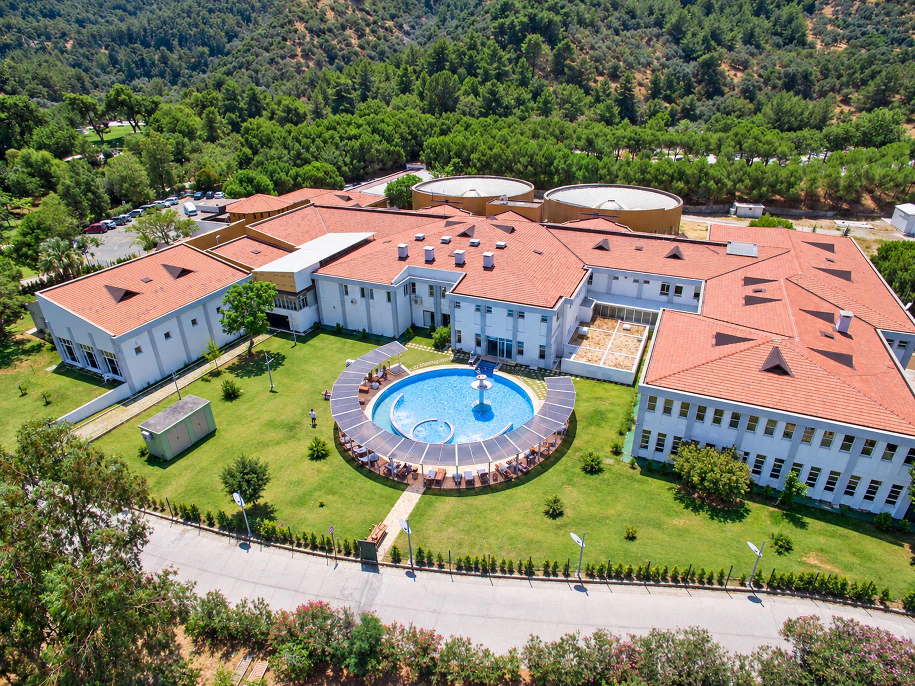 A large building with a central circular pool, surrounded by multiple smaller buildings and a parking area.