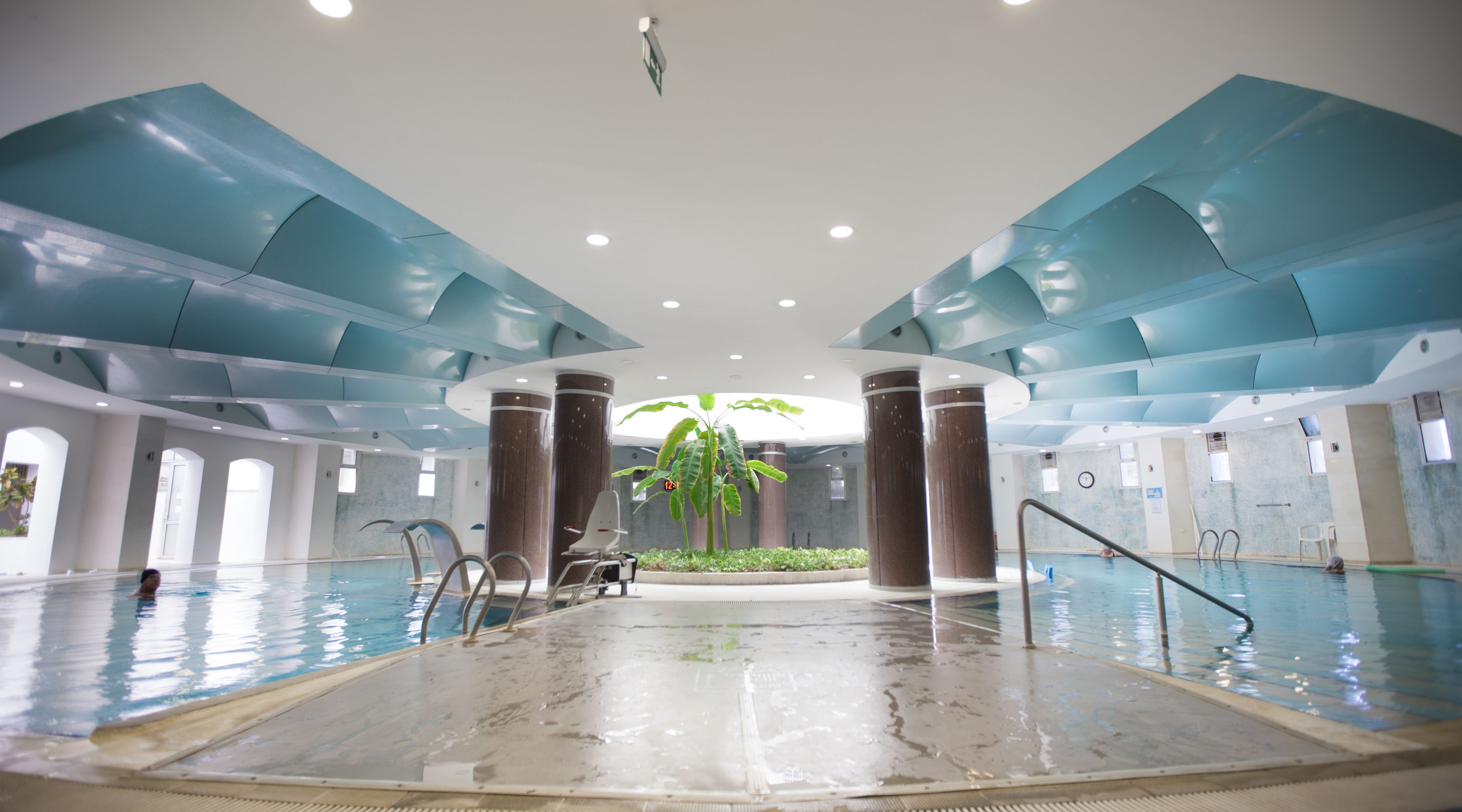 Uma grande piscina interior com uma ilha central, rodeada por colunas e um teto com painéis azuis.