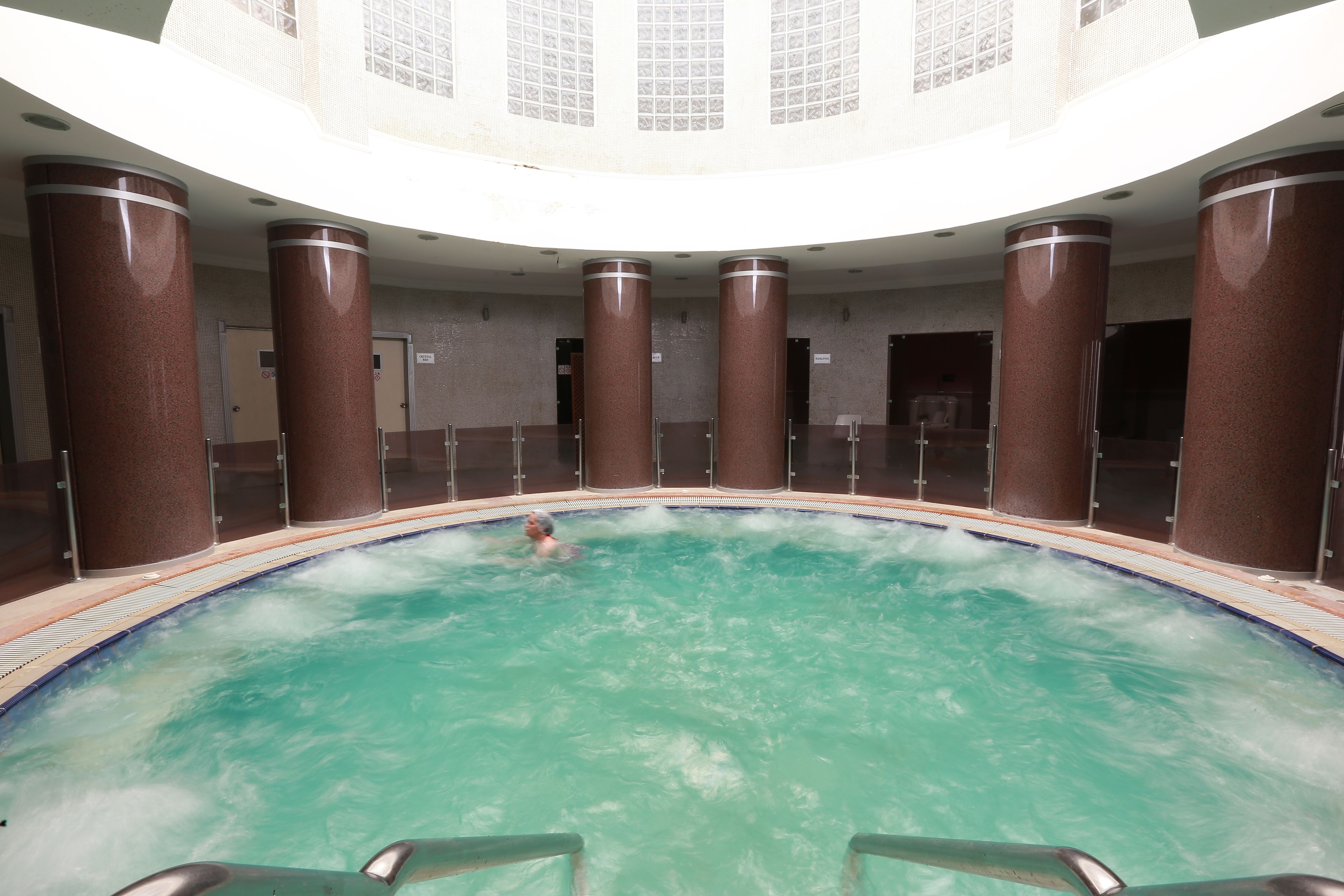 A large indoor swimming pool with a person swimming, surrounded by tall pillars and a high ceiling with a patterned design.