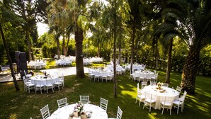 An outdoor wedding venue with round tables and white chairs, surrounded by lush greenery and palm trees. - Balcova Thermal Hotel (Izmir)