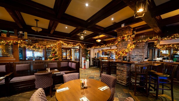 Un ristorante con travi in legno, pareti in mattoni e decorazioni festive. Si notano tavoli rotondi con sedie, una zona bar e un'area salotto con panche imbottite.