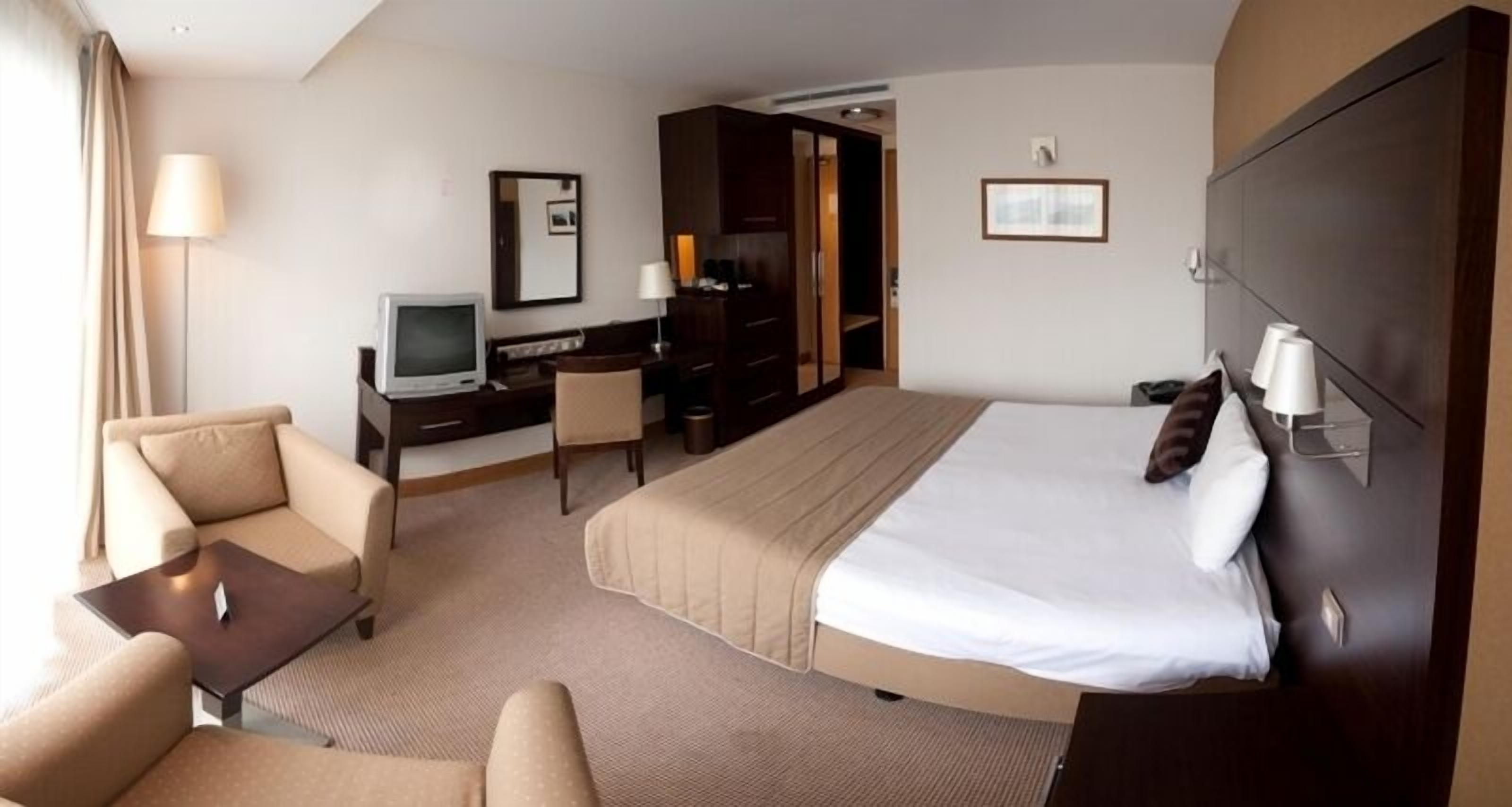 A hotel room with a bed, a desk, a chair, a television, and a lamp.