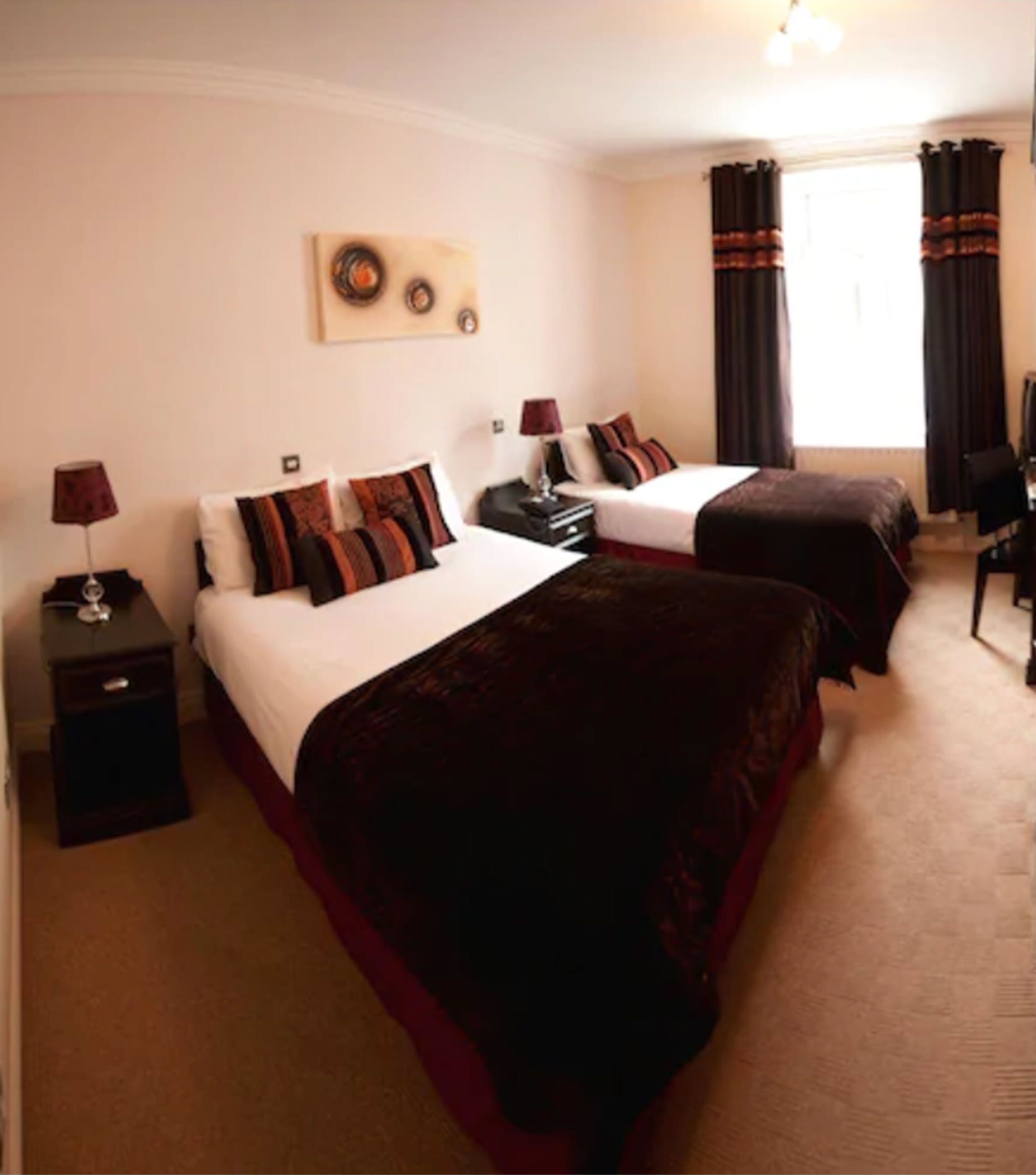 A hotel room with two beds, each with a red and white bedspread, a nightstand with a lamp, and a wall-mounted artwork above the beds.
