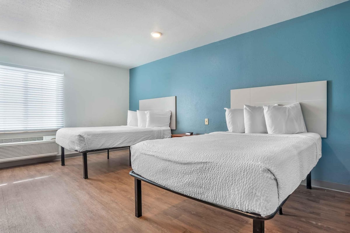 A hotel room with two beds, a window with blinds, a ceiling light, and a blue wall.