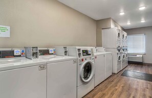 A laundry facility with multiple washing machines and dryers, a sign indicating products purchased at the desk, and a window with blinds.