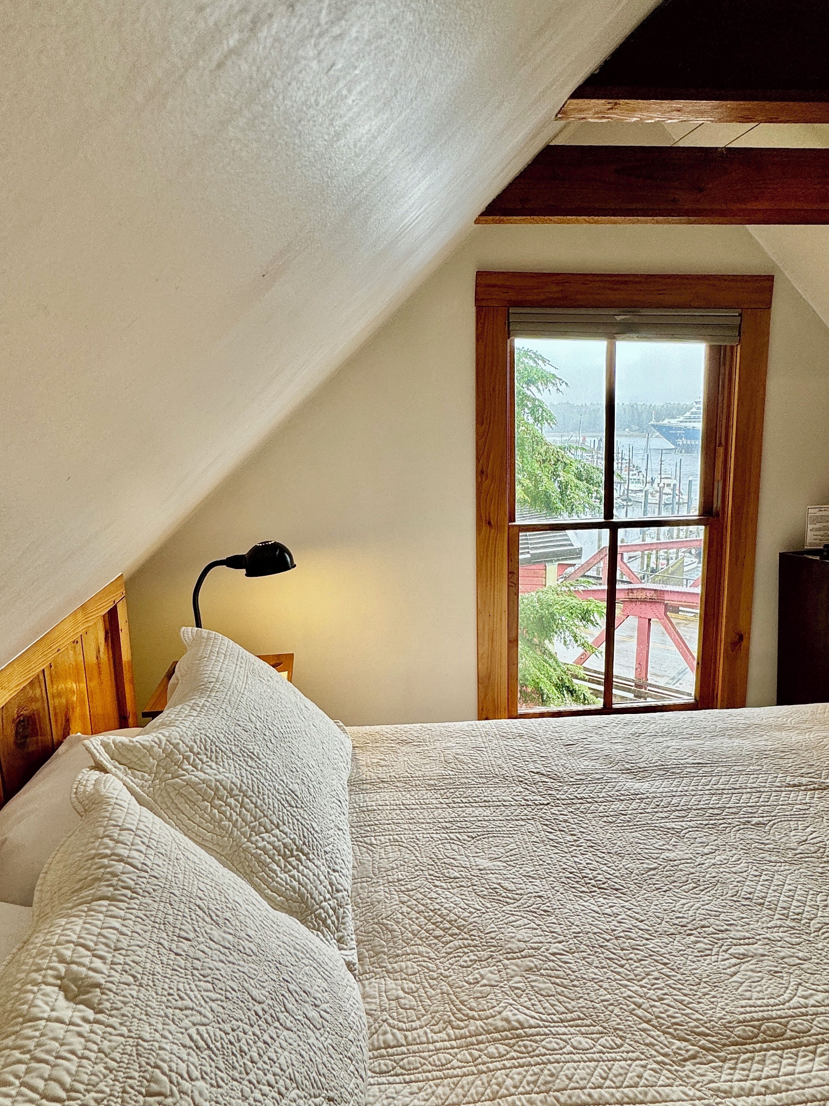 A bedroom with a sloped ceiling, a wooden headboard, a large bed with white bedding, a nightstand with a lamp, and a window with a view of a red bridge and greenery.
