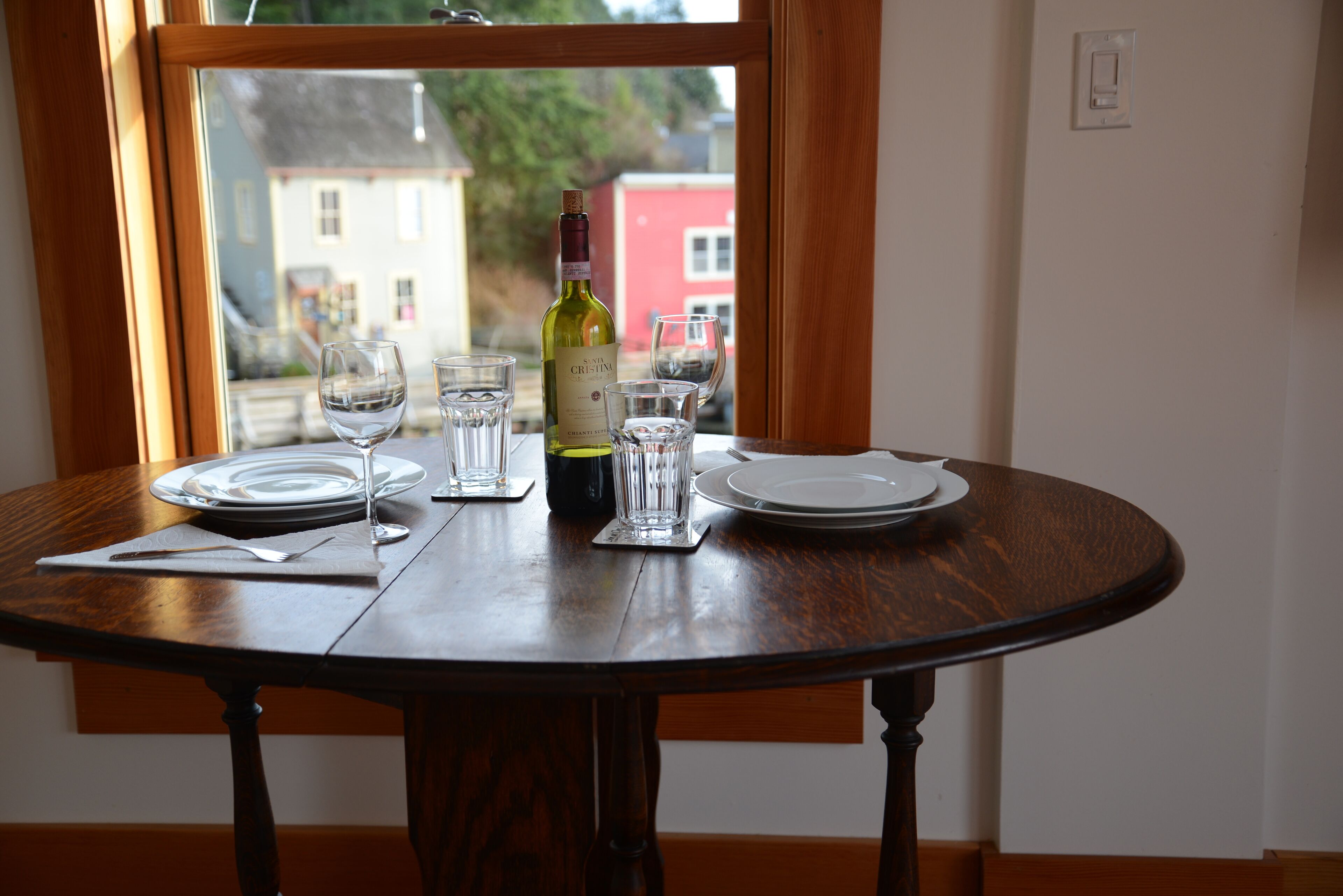 Una mesa de comedor preparada con platos, vasos y una botella de vino, ubicada junto a una ventana con vista a edificios.
