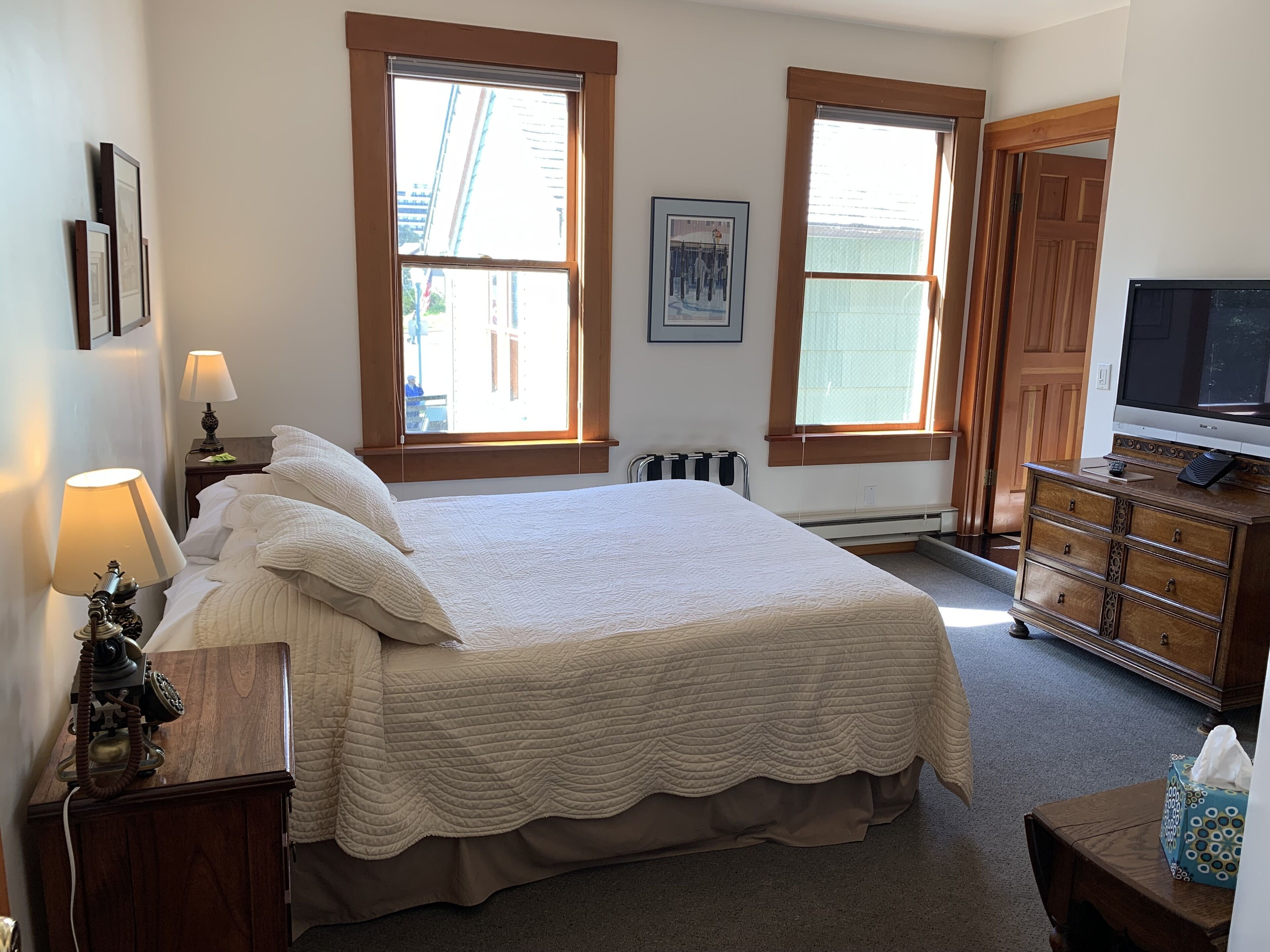 A bedroom with a large bed, two bedside lamps, a wooden dresser, a television, and two windows with blinds.
