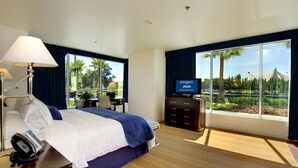 A hotel room with a bed, a desk with a computer, a TV, and large windows with a view of greenery.