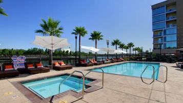 A rooftop pool area with lounge chairs and umbrellas, surrounded by palm trees and a high-rise building.