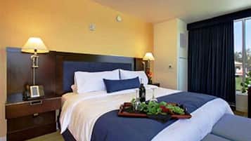 Habitación de hotel con una cama grande, dos mesitas de noche con lámparas, una bandeja con frutas y una botella sobre la cama, y una ventana con cortinas.