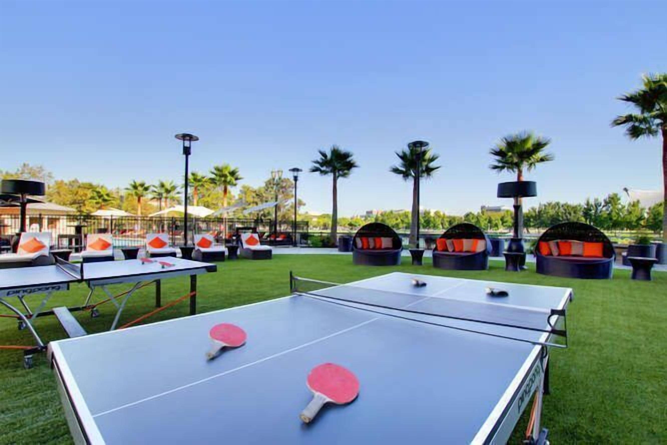 Zona de ping pong al aire libre con mesas y sillas, rodeada de palmeras y un cielo despejado.