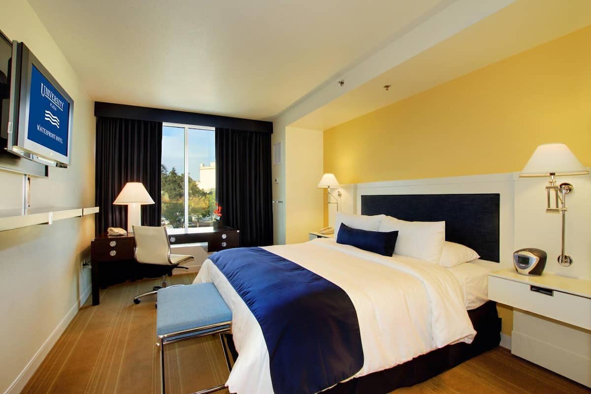 A hotel room with a bed, bedside tables, a desk, a chair, a TV, and a window with curtains.