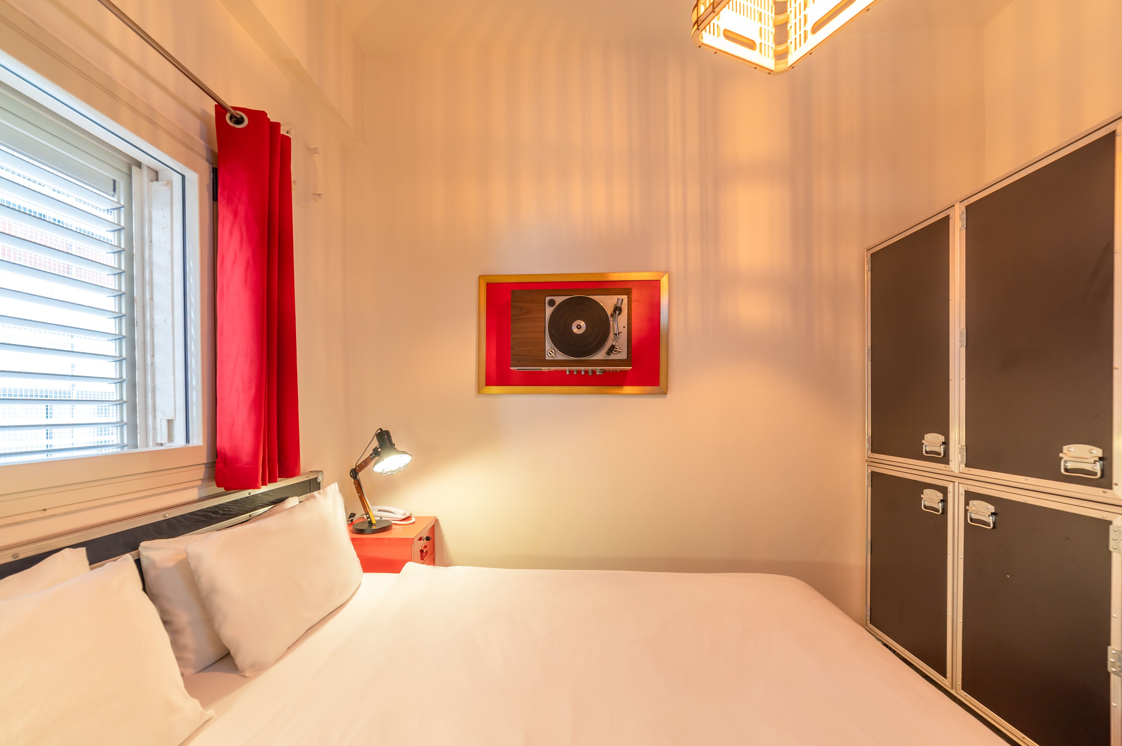Ett hotellrum med en säng, ett skrivbord med en lampa, en garderob, ett fönster med persienner och en tavla som hänger på väggen.
