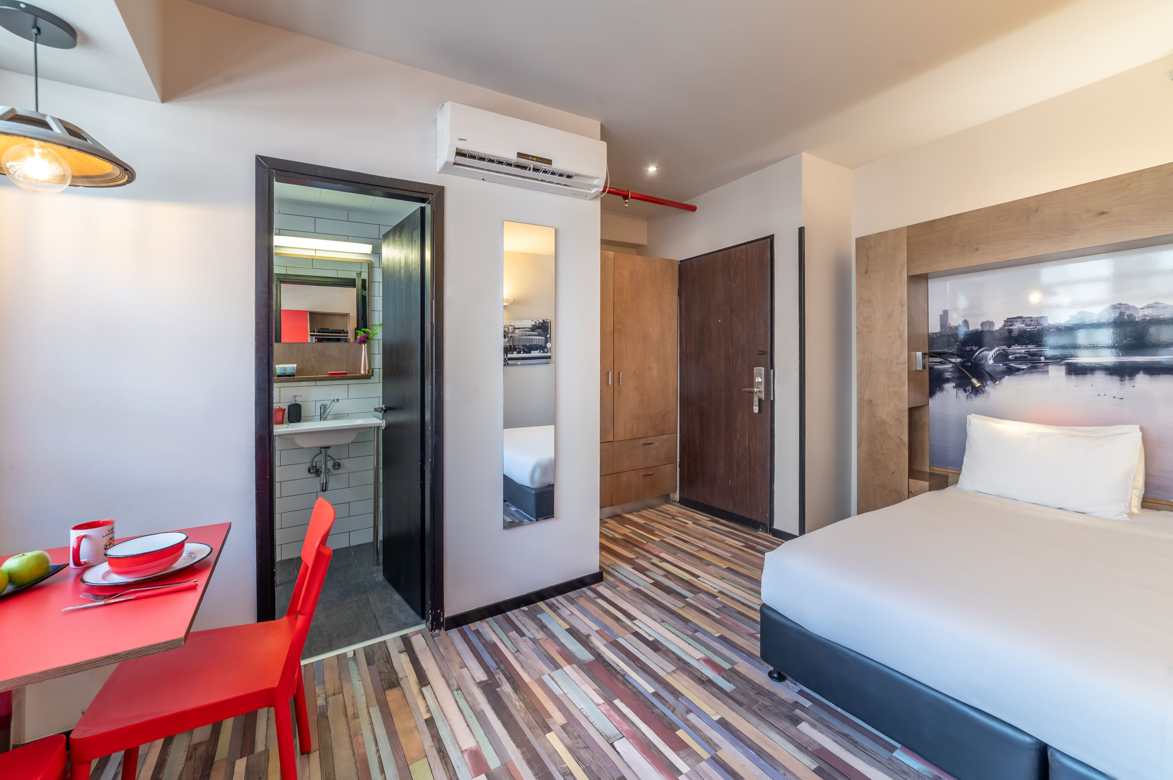 A modern hotel room with a bed, a dining table with red chairs, a kitchen area, and a large window with a cityscape view.
