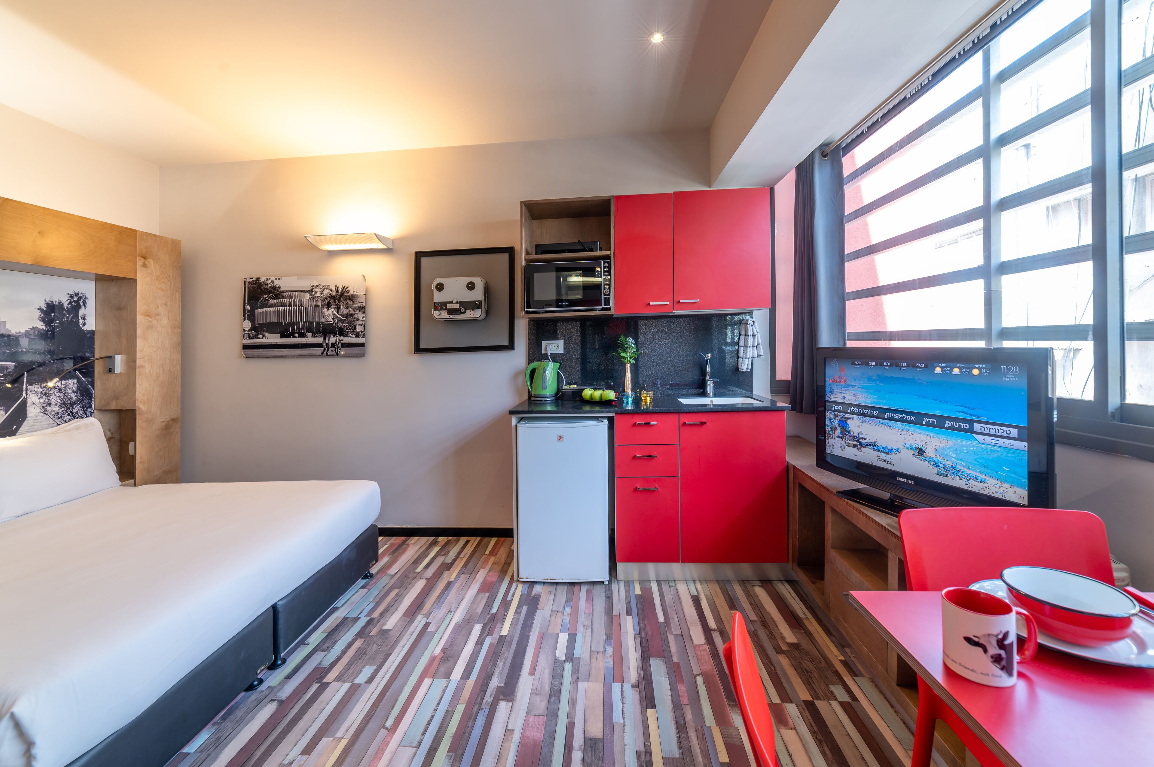A compact hotel room with a bed, kitchenette, and a TV displaying a beach scene.