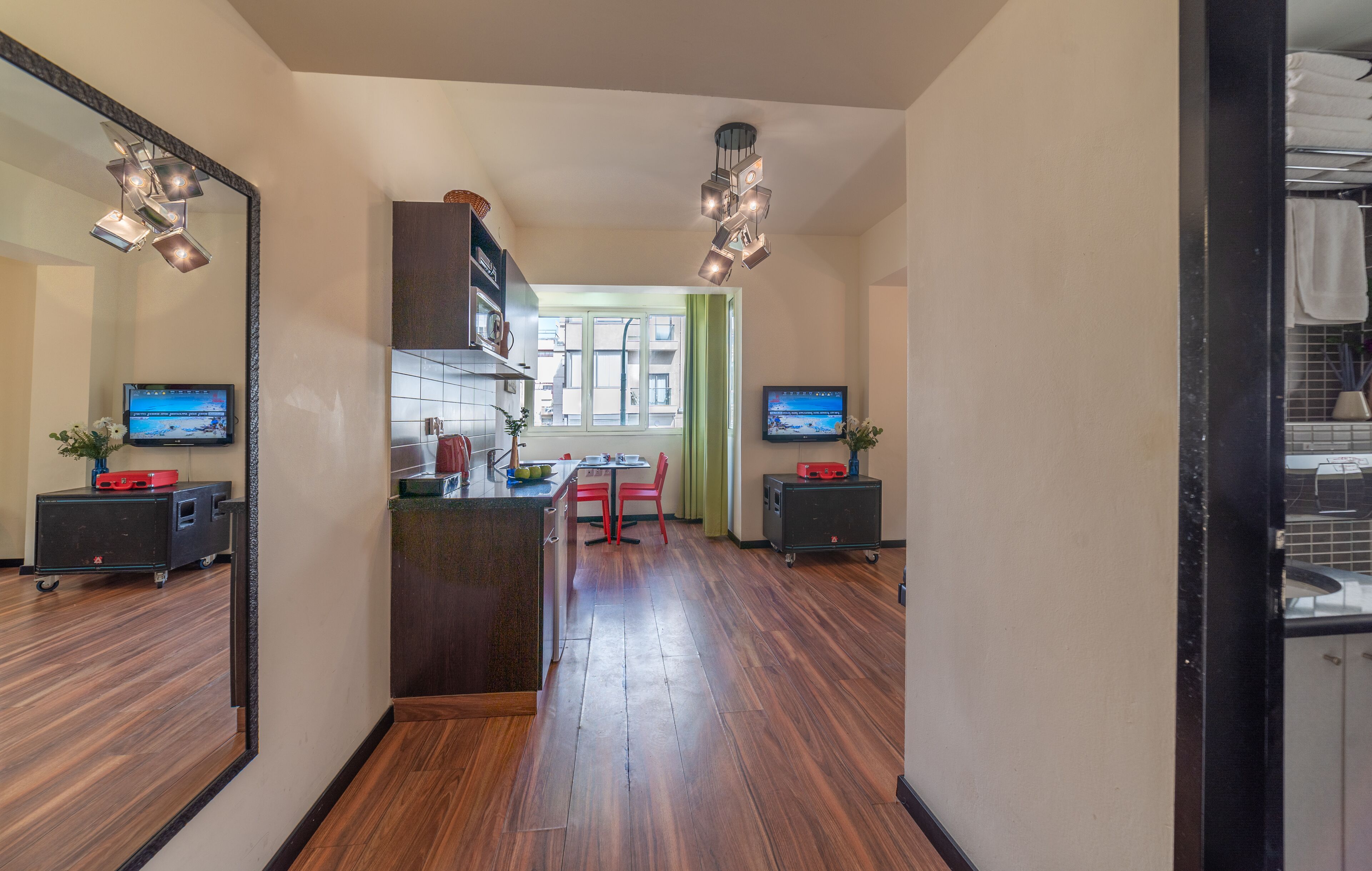 A modern hotel room with a wooden floor, a kitchen area with a dining table and chairs, and two TVs mounted on the walls.