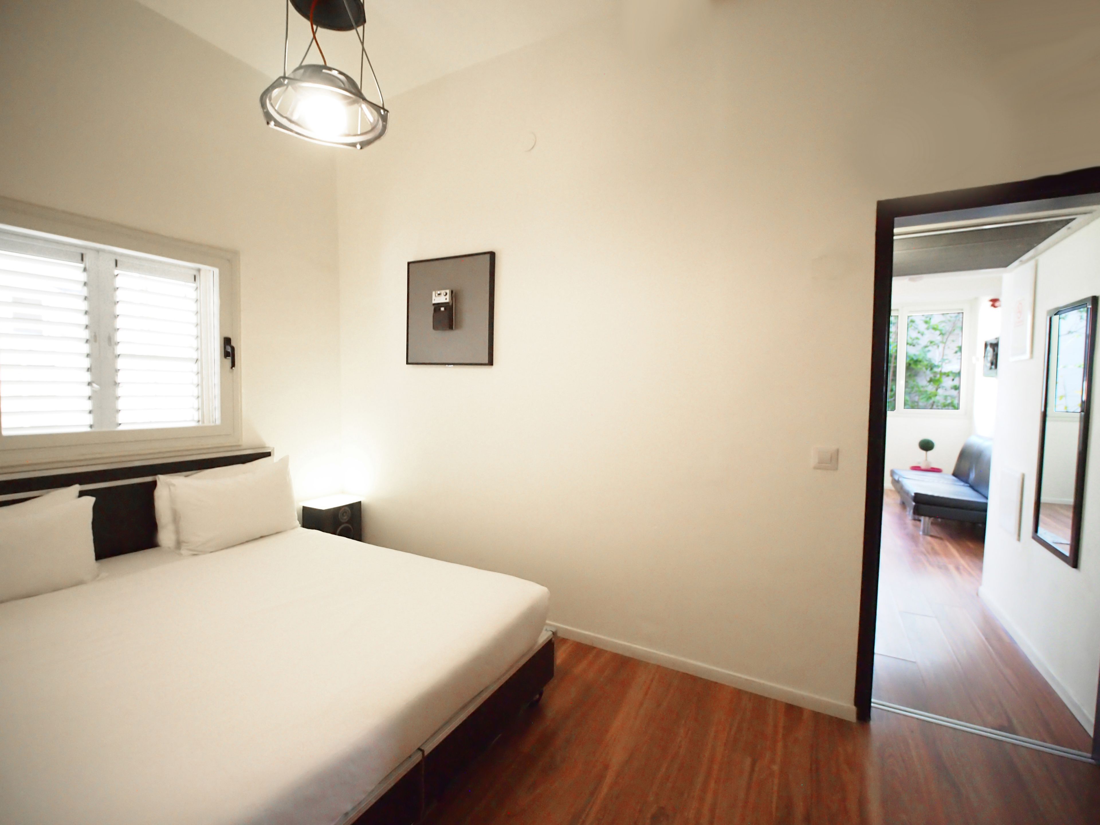 A bedroom with a bed, a bedside table, a wall-mounted light, a window with blinds, and a door leading to another room.