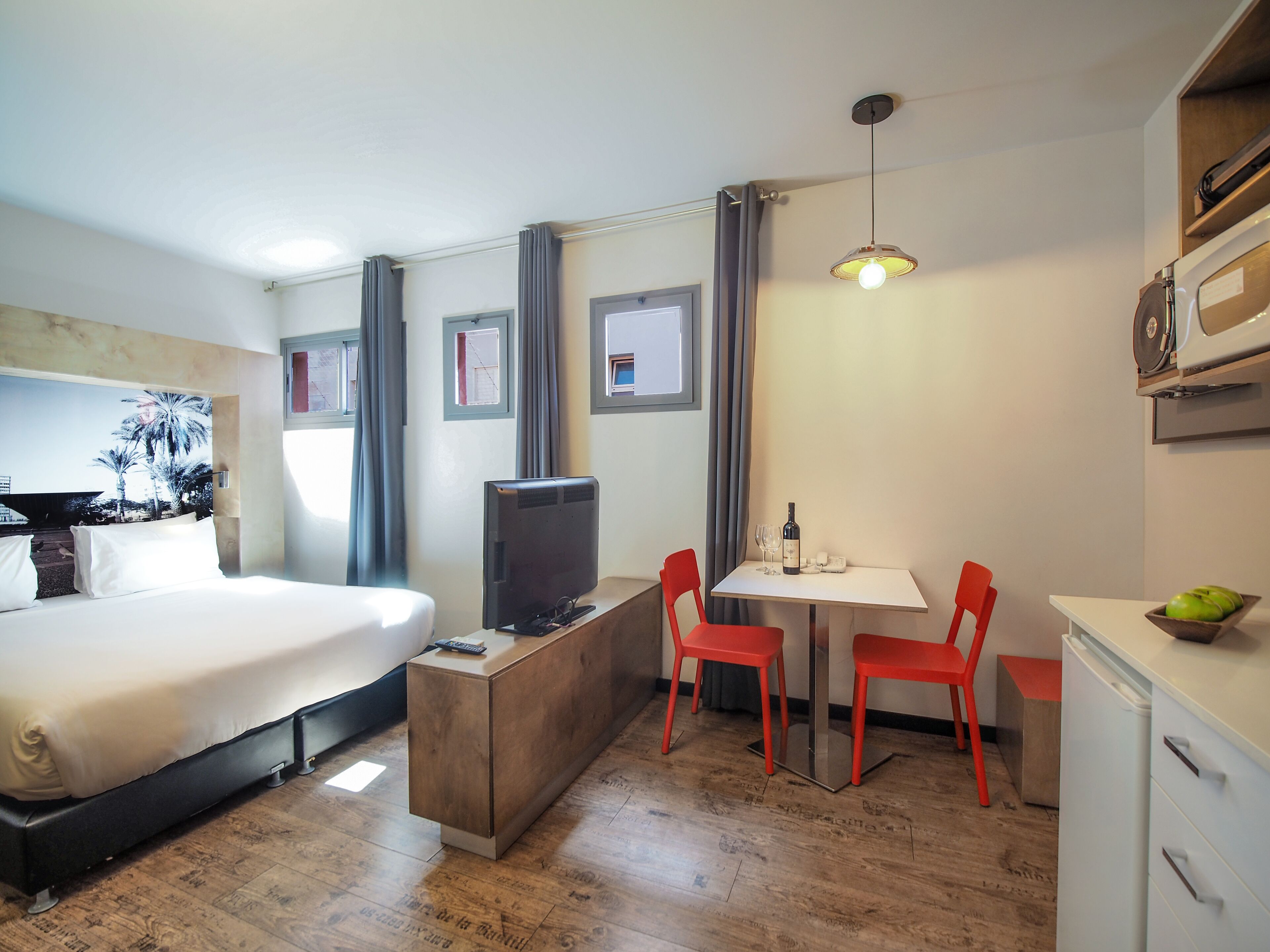 A hotel room with a bed, a desk with a television, a dining table with chairs, and a small refrigerator.