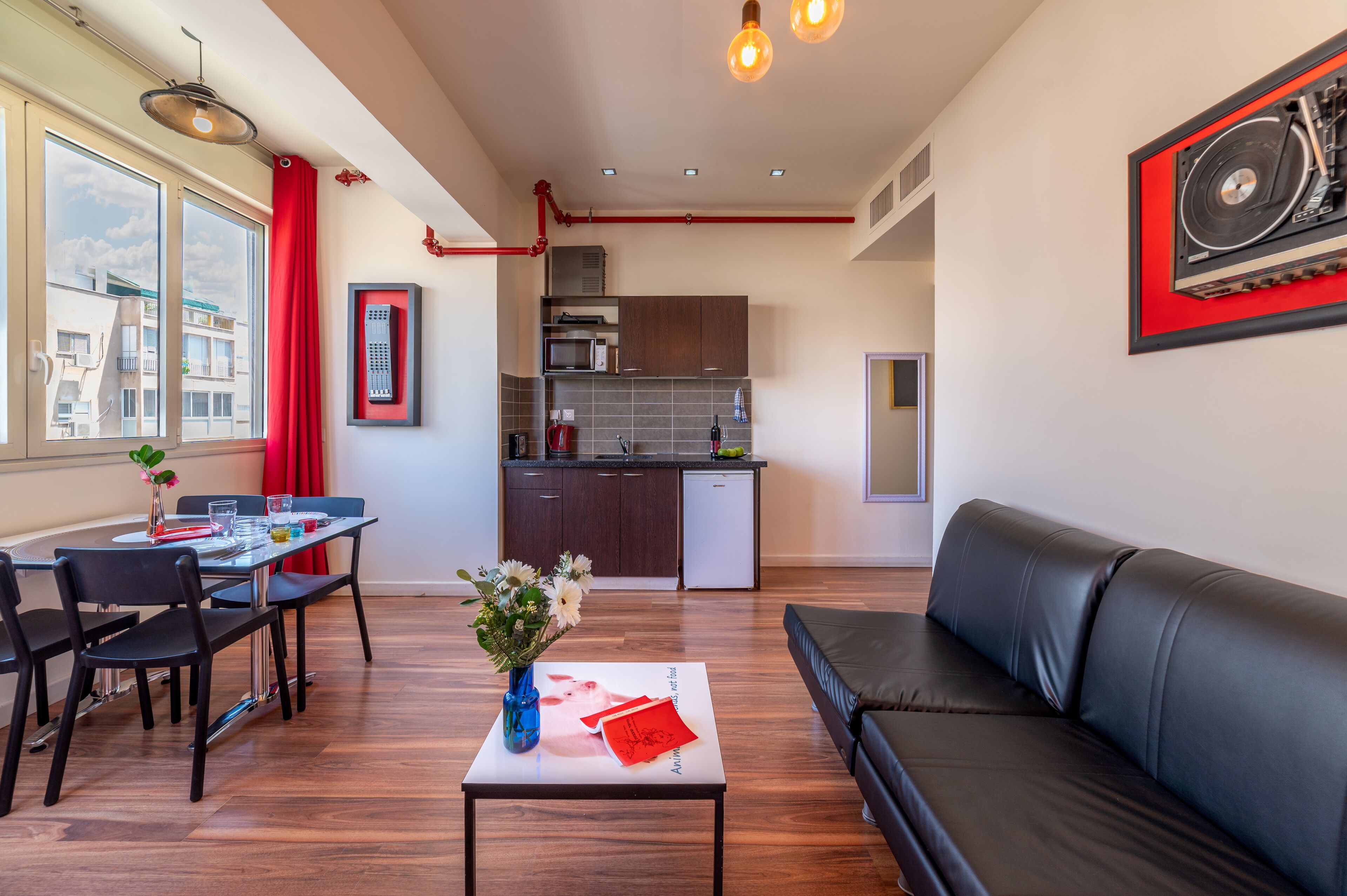 A modern living room with a black leather sofa, a dining area with a table and chairs, a kitchen with dark cabinets and a microwave, and a large window with red curtains.