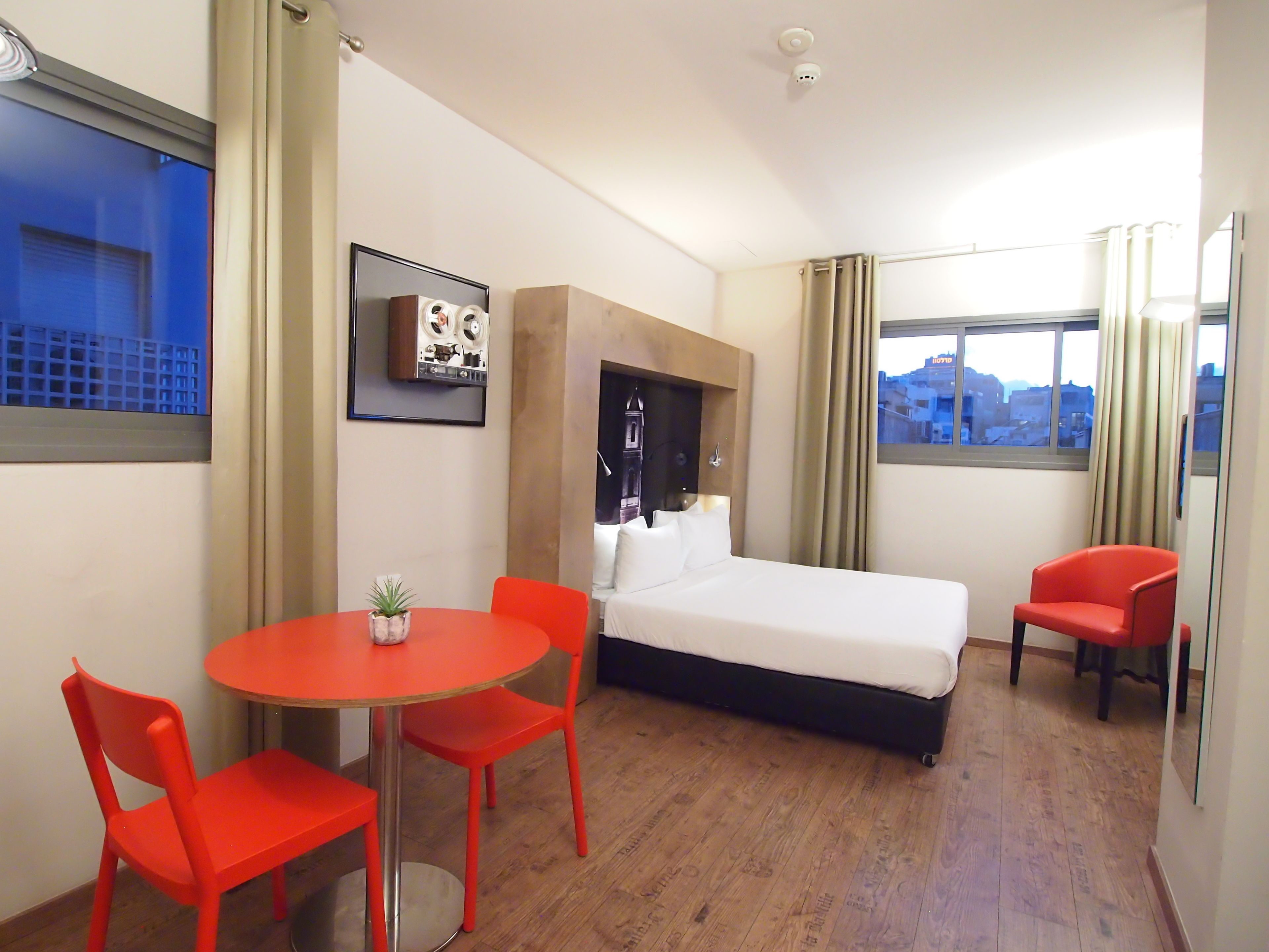 A hotel room with a bed, a desk with a chair, a small table with a plant, and a window with curtains.