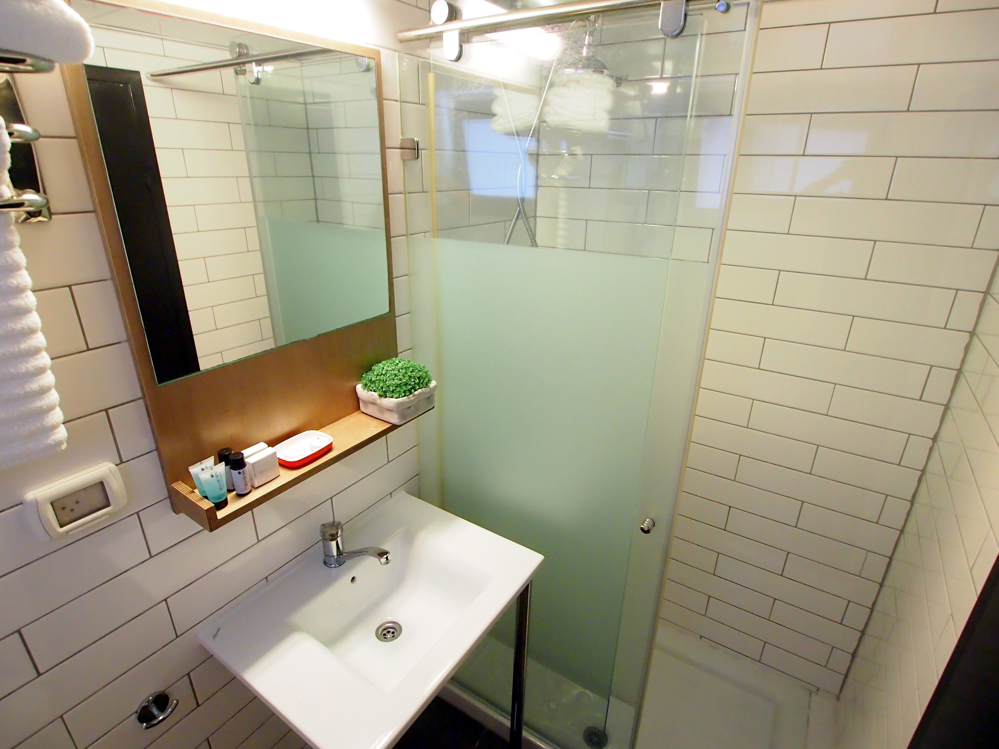A bathroom with a white sink, a mirror, a glass shower enclosure, and a small shelf with toiletries.
