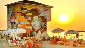 A beachside building with colorful murals, a digital clock, and beach chairs and umbrellas. - Ben Yehuda Apartments (Tel Aviv)