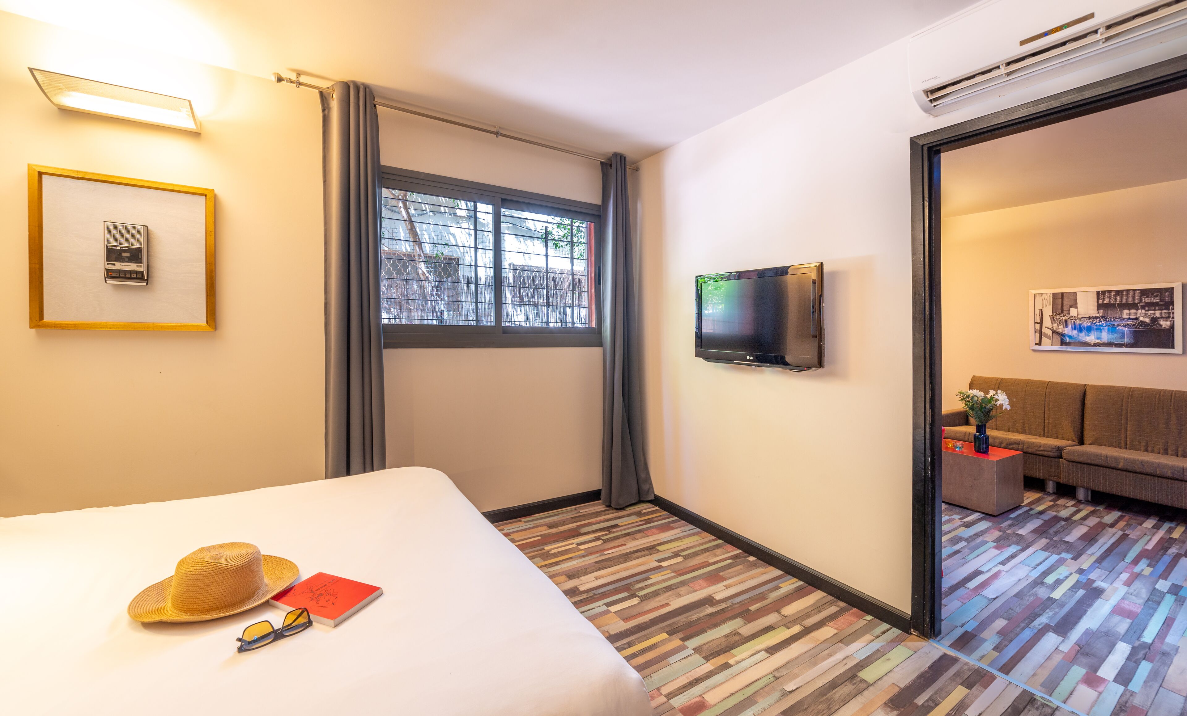A hotel room with a bed, a TV, a window with curtains, a framed picture, and a colorful wooden floor.