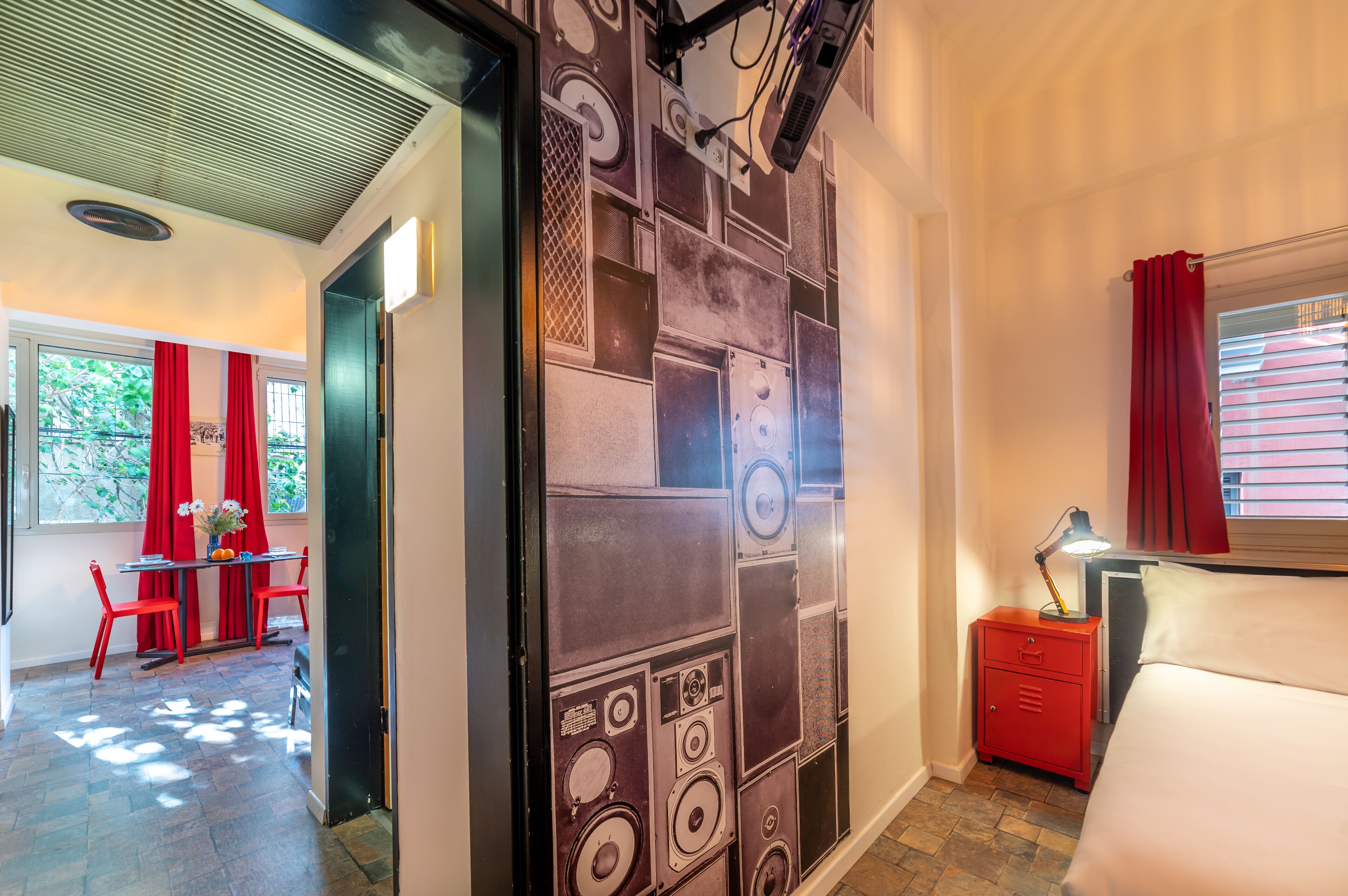 Una habitación con una cama, una mesita de noche con una lámpara, una silla roja, una mesa con un florero y una pared con un diseño original.