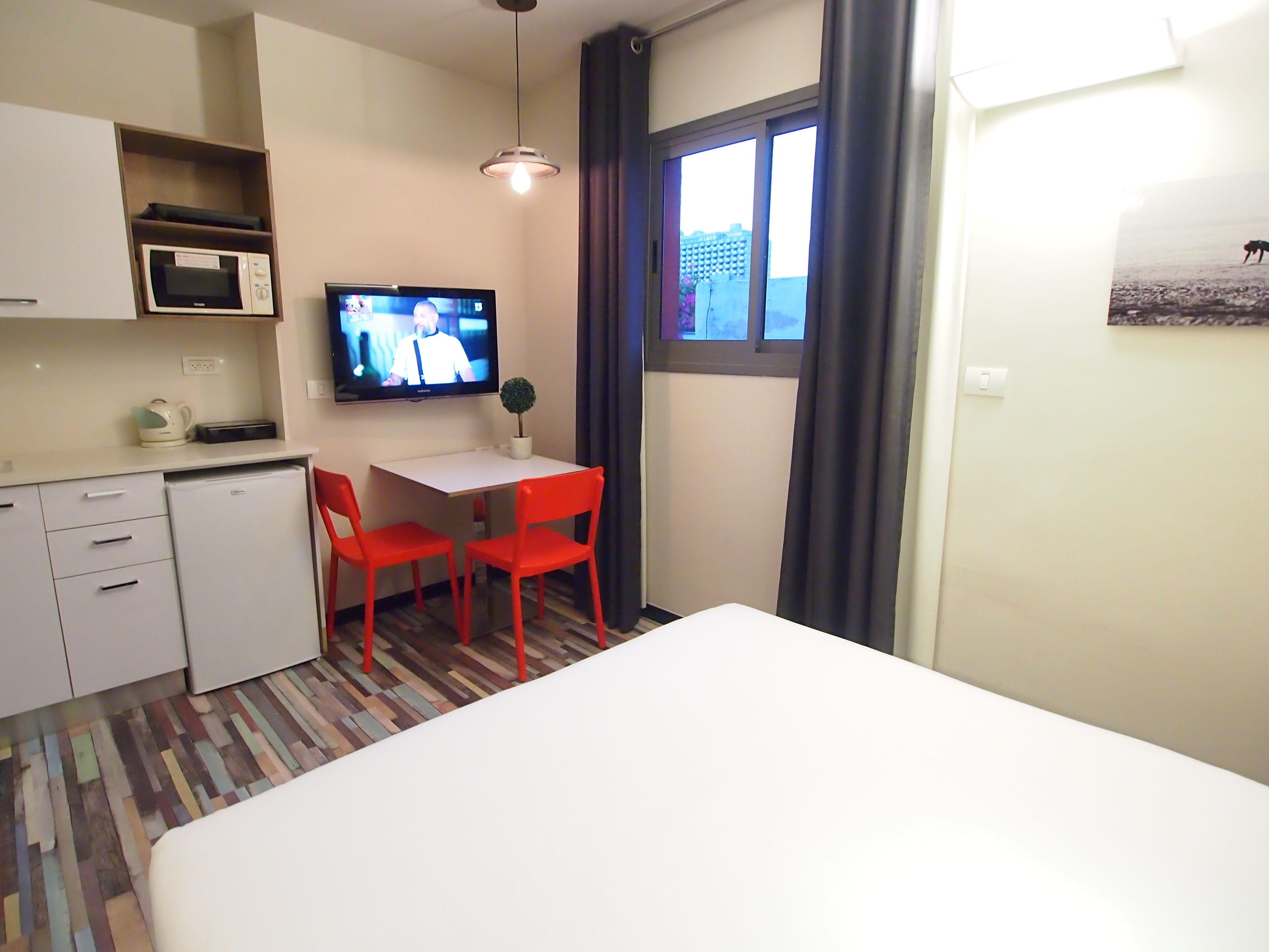 Una habitación de hotel compacta con cama, una kitchenette pequeña, un área de comedor con mesa y sillas, televisor y ventana con cortinas.