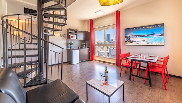 A modern living room with a black spiral staircase, a dining area with red chairs, and a kitchen in the background. - Ben Yehuda Apartments (Tel Aviv)