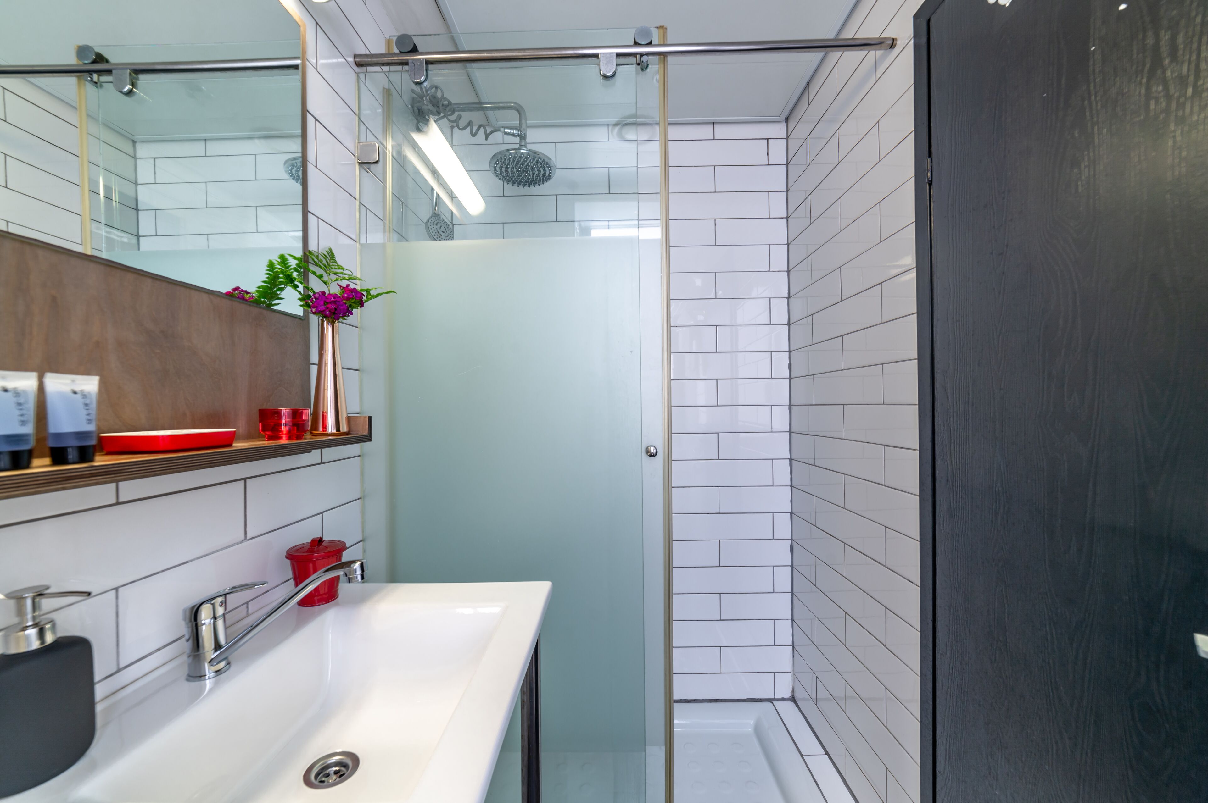 A modern bathroom with a glass shower enclosure, a white sink, and a wooden shelf with toiletries.