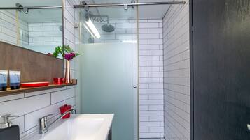 A modern bathroom with a glass shower enclosure, a white sink, and a wooden shelf with toiletries.