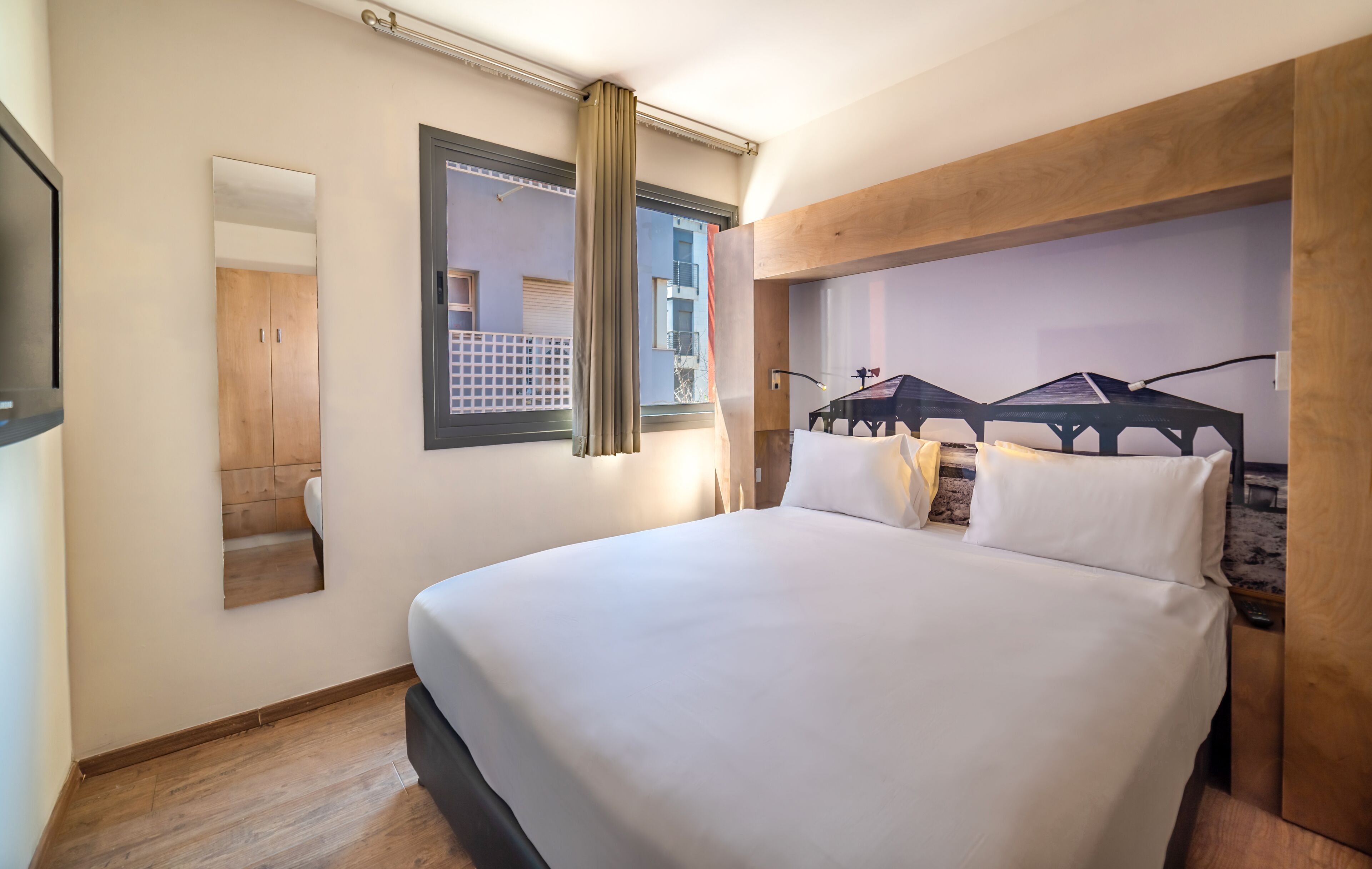 Une chambre d’hôtel avec un grand lit, un miroir, une fenêtre avec des rideaux et une tête de lit en bois.