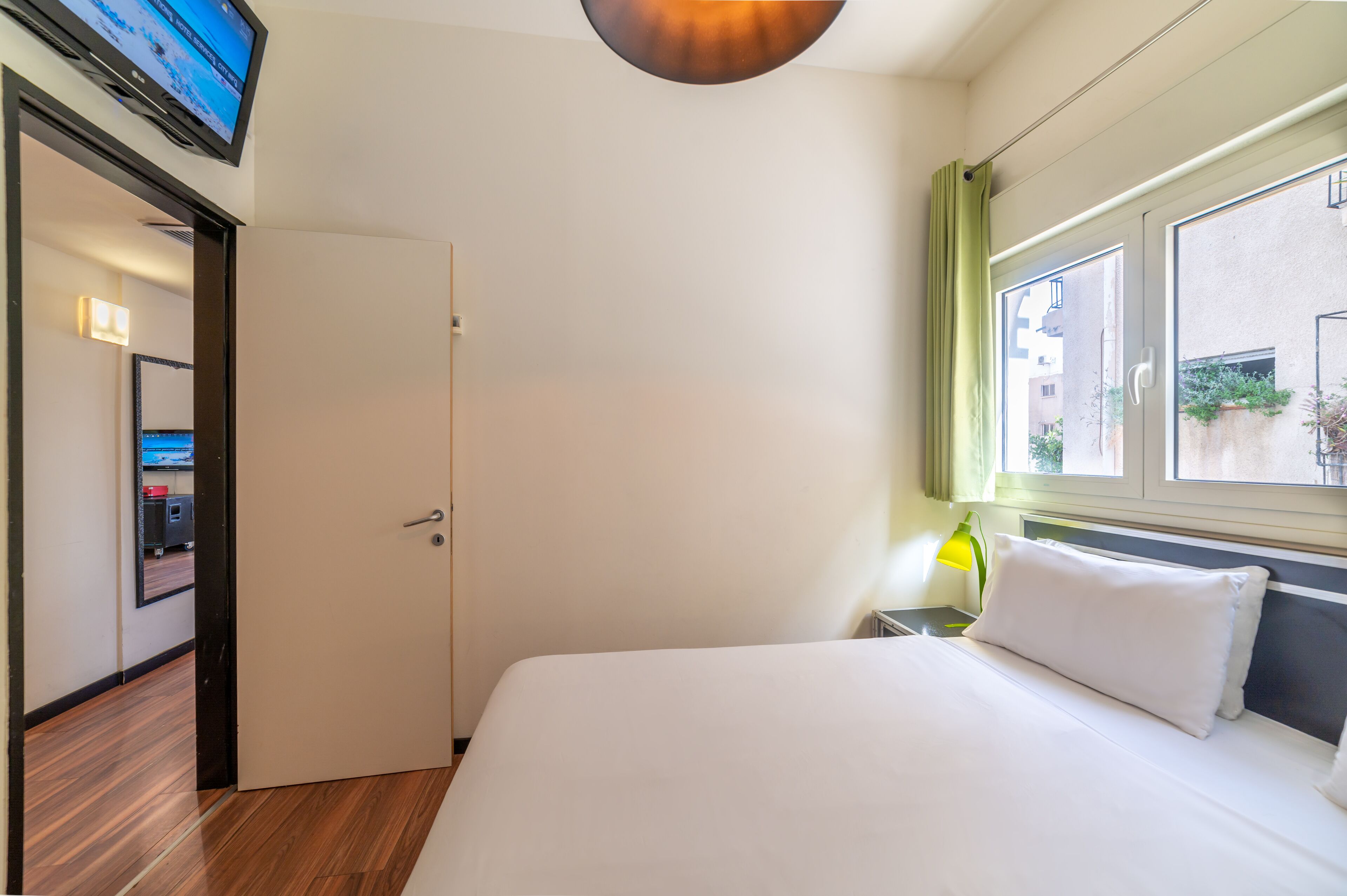 Ett hotellrum med en säng, en tv som är monterad på väggen, ett fönster med utsikt över byggnader och en dörr som leder till ett annat rum.