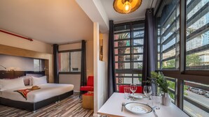 A modern hotel room with a bed, a dining table set with glasses and cutlery, and large windows offering a view of the street. - Ben Yehuda Apartments (Tel Aviv)